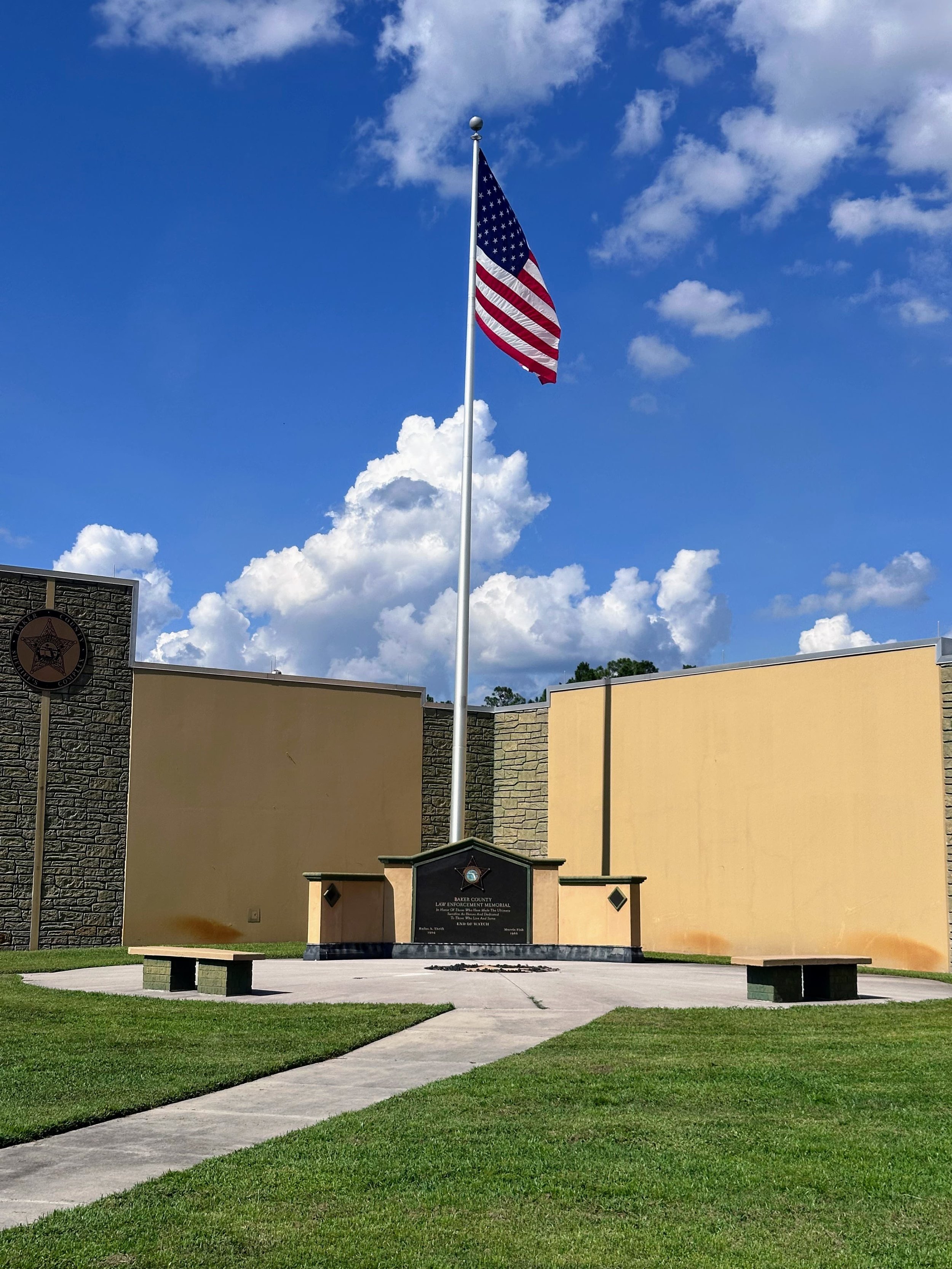A flagpole with the American flag flying in front of a memorial monument, with a blue sky and scattered clouds overhead. The memorial is surrounded by a paved area with benches and green grass.