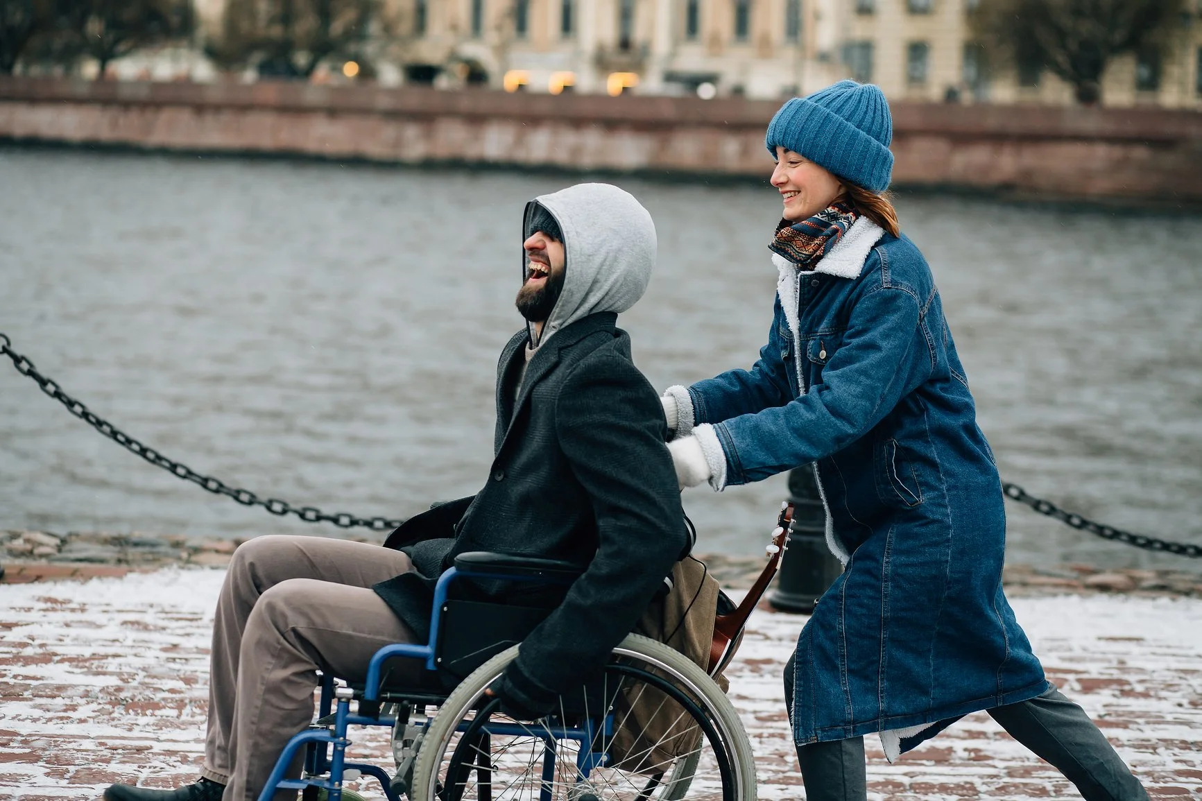 Man sitting in wheelchair, being pushed by a woman. They are wearing coats and beanies and laughing.