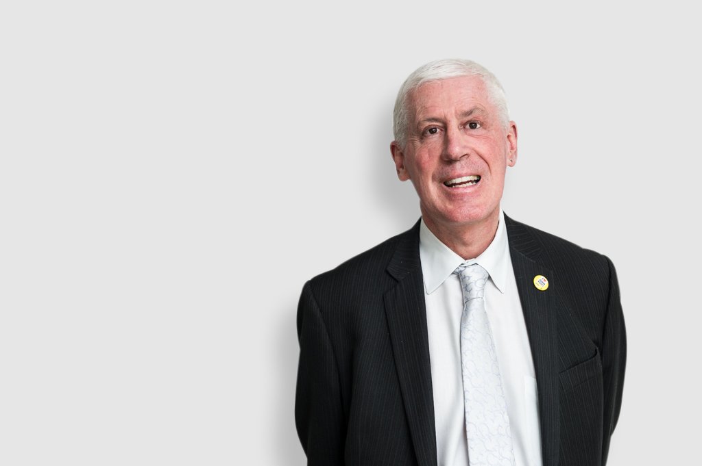 Graeme Innes stands in front of a white wall. He is wearing a black suit with a white shirt and silver tie. He has grey hair and is smiling at the camera.