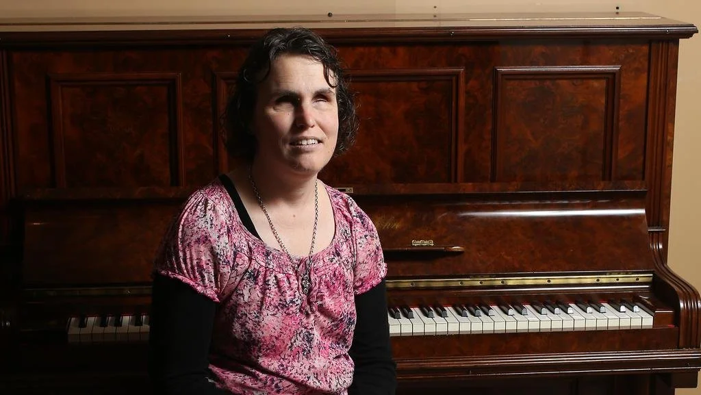 Emma Bennison sits at a wooden upright piano. Her eyes are closed and she has chin-length brown wavy hair. She is wearing a pink patterned top and a silver necklace.