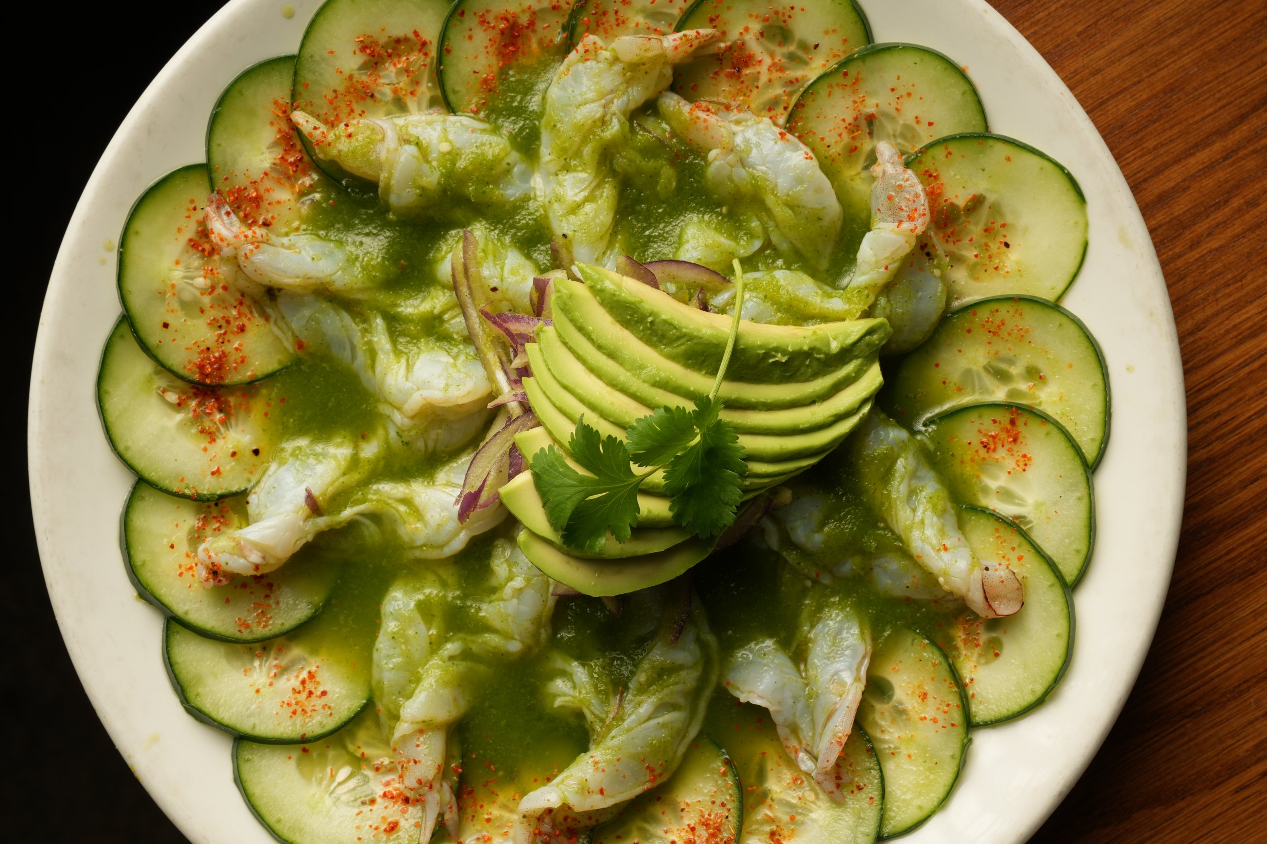 A plate of shrimp ceviche garnished with sliced avocado, cucumber, cilantro, and red seasoning, with a green sauce drizzle.