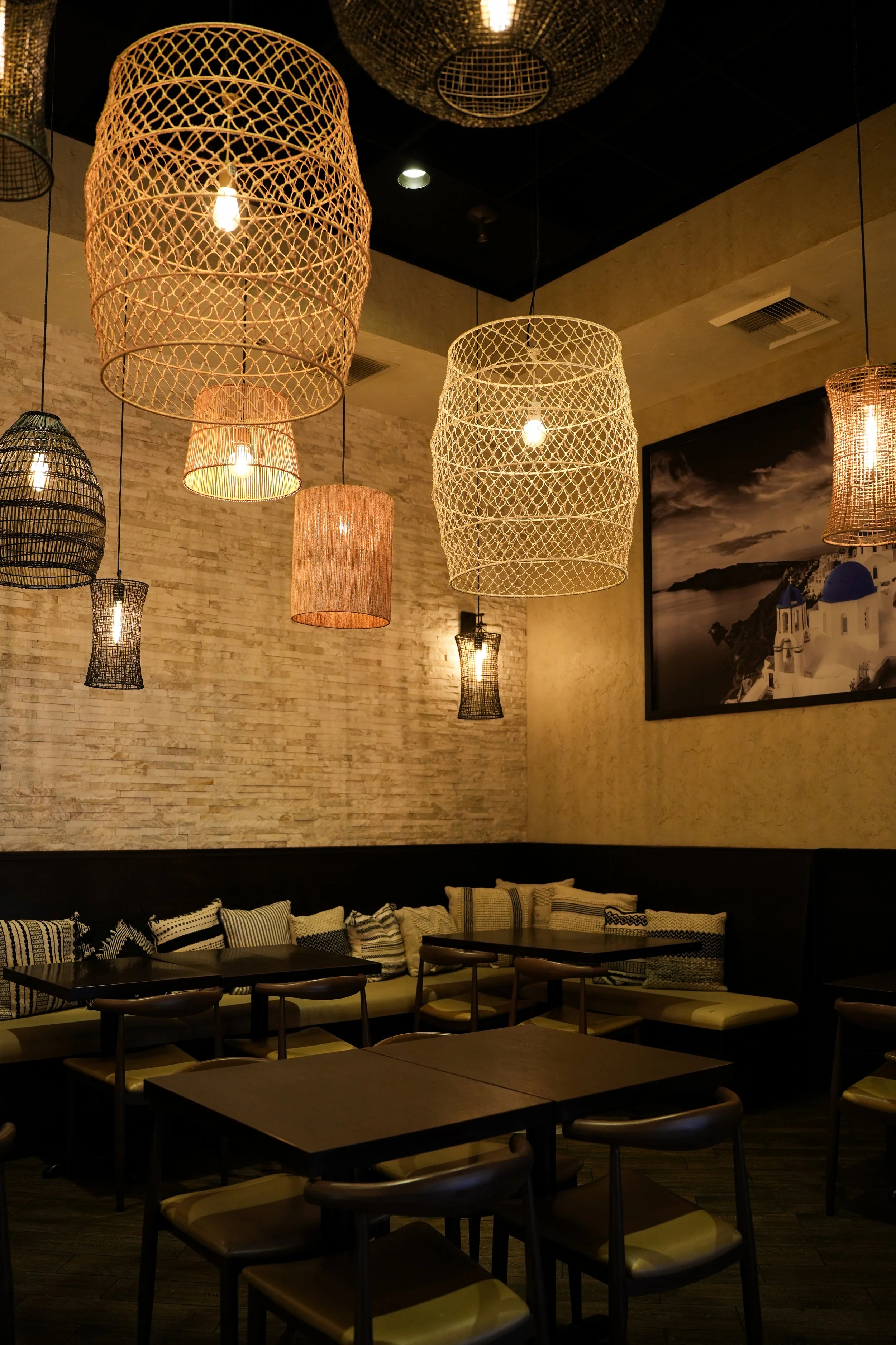 Dimly lit restaurant or cafe with hanging woven light fixtures and a row of tables with cushioned chairs and pillows, beige brick wall, and a large framed black and white photo of a landscape.