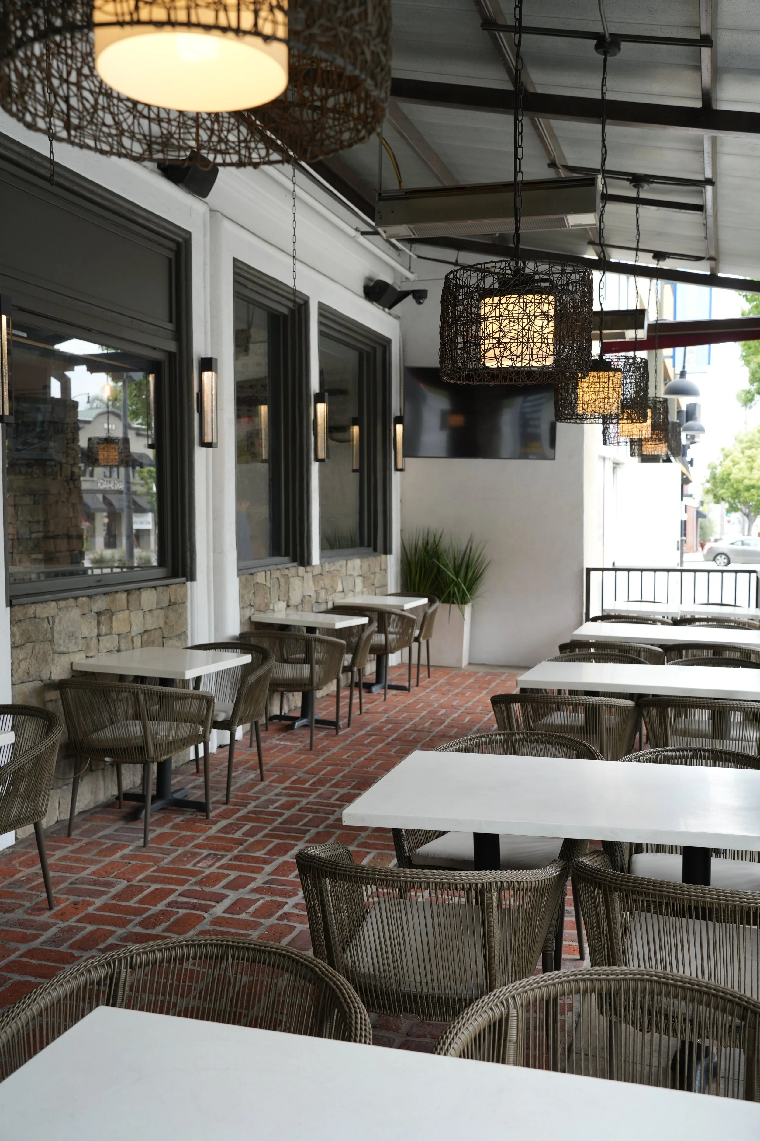 Empty outdoor patio restaurant or cafe with white tables, woven chairs, brick floor, and modern hanging light fixtures.