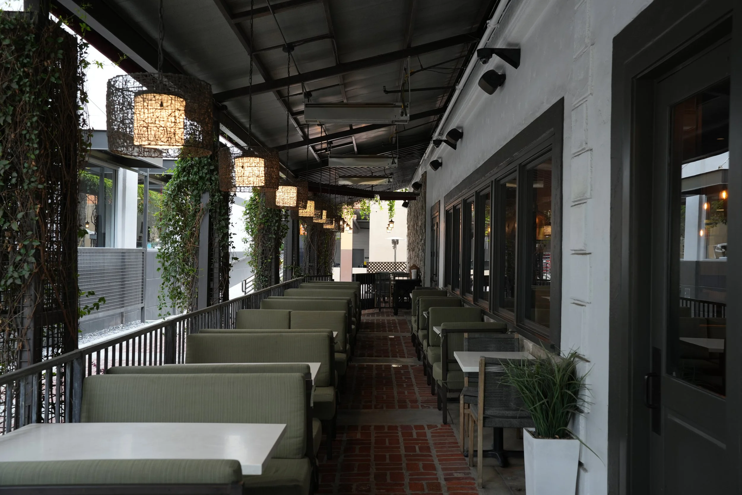 Empty outdoor patio with green upholstered chairs, white tables, potted plants, hanging lights, and large windows.