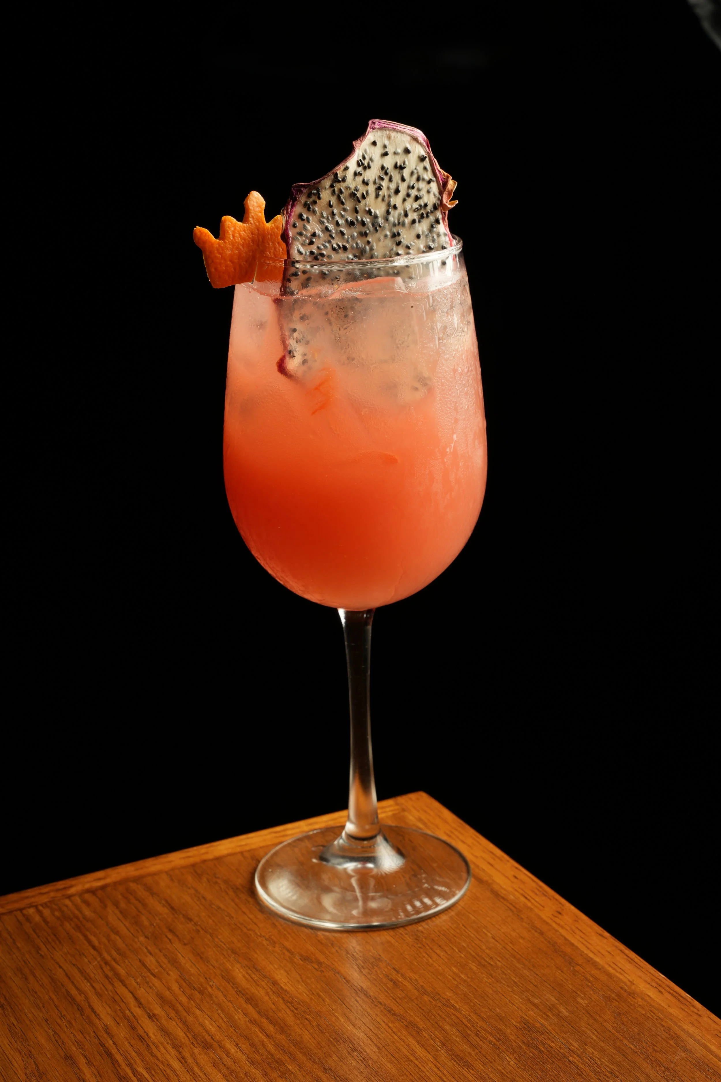 A pink cocktail in a stemmed glass garnished with a piece of dragon fruit and a small orange peel layered with a crown-shaped decoration, set on a wooden surface against a black background.