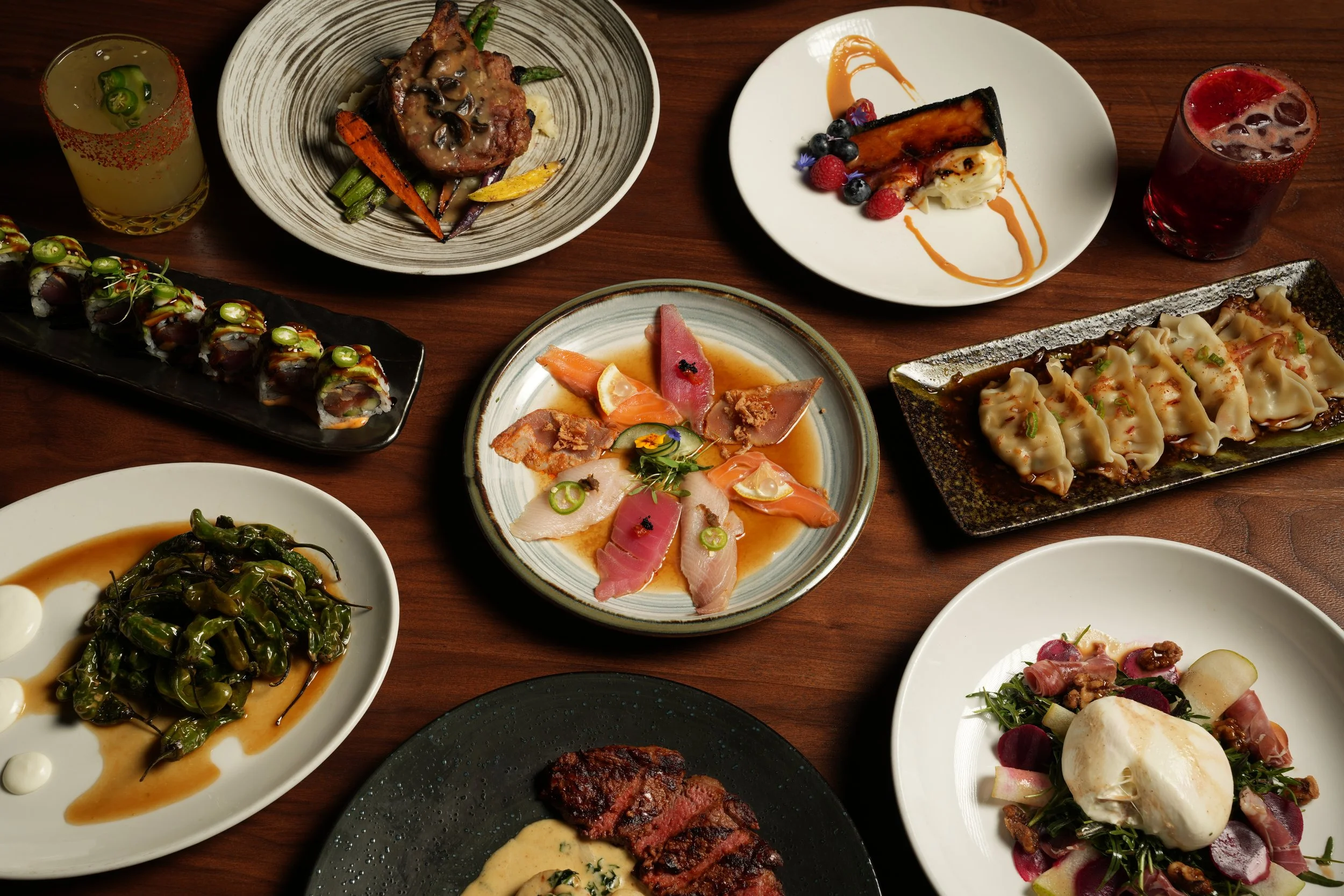 Assorted dishes of sushi, grilled meats, salads, and desserts on a wooden table with drinks.