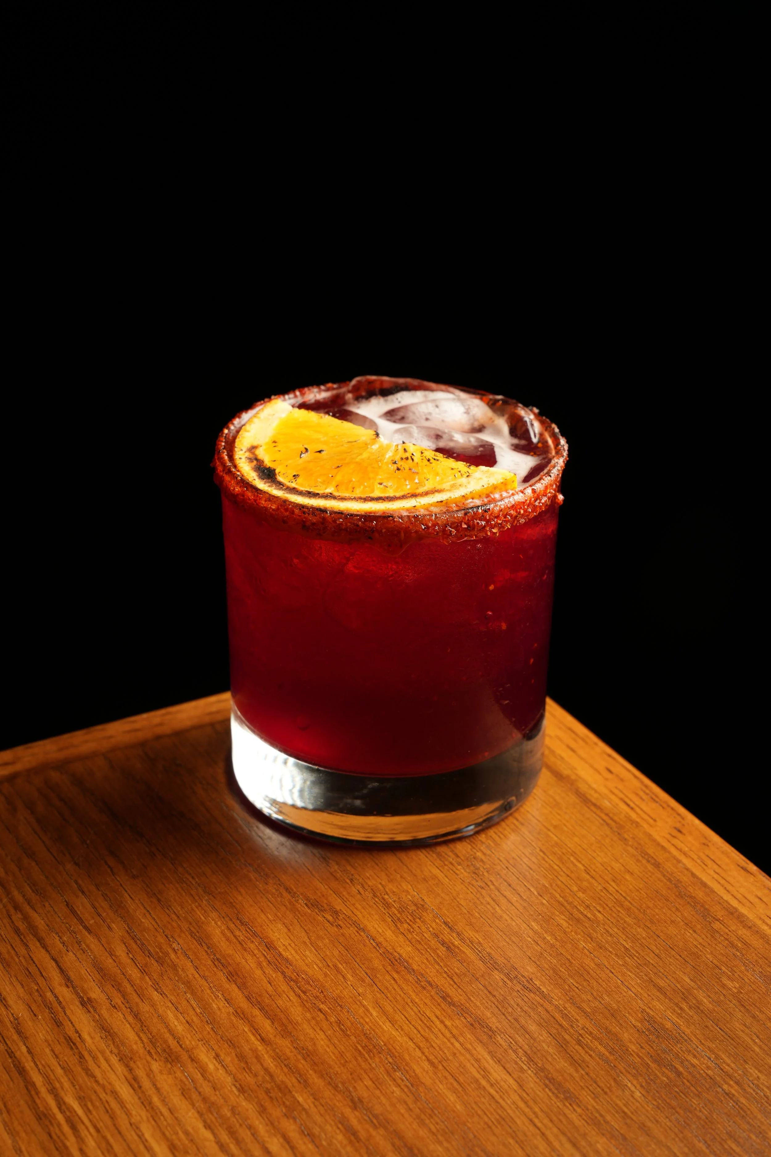 A red cocktail drink in a clear glass, garnished with a slice of orange, set on a wooden surface with a black background.