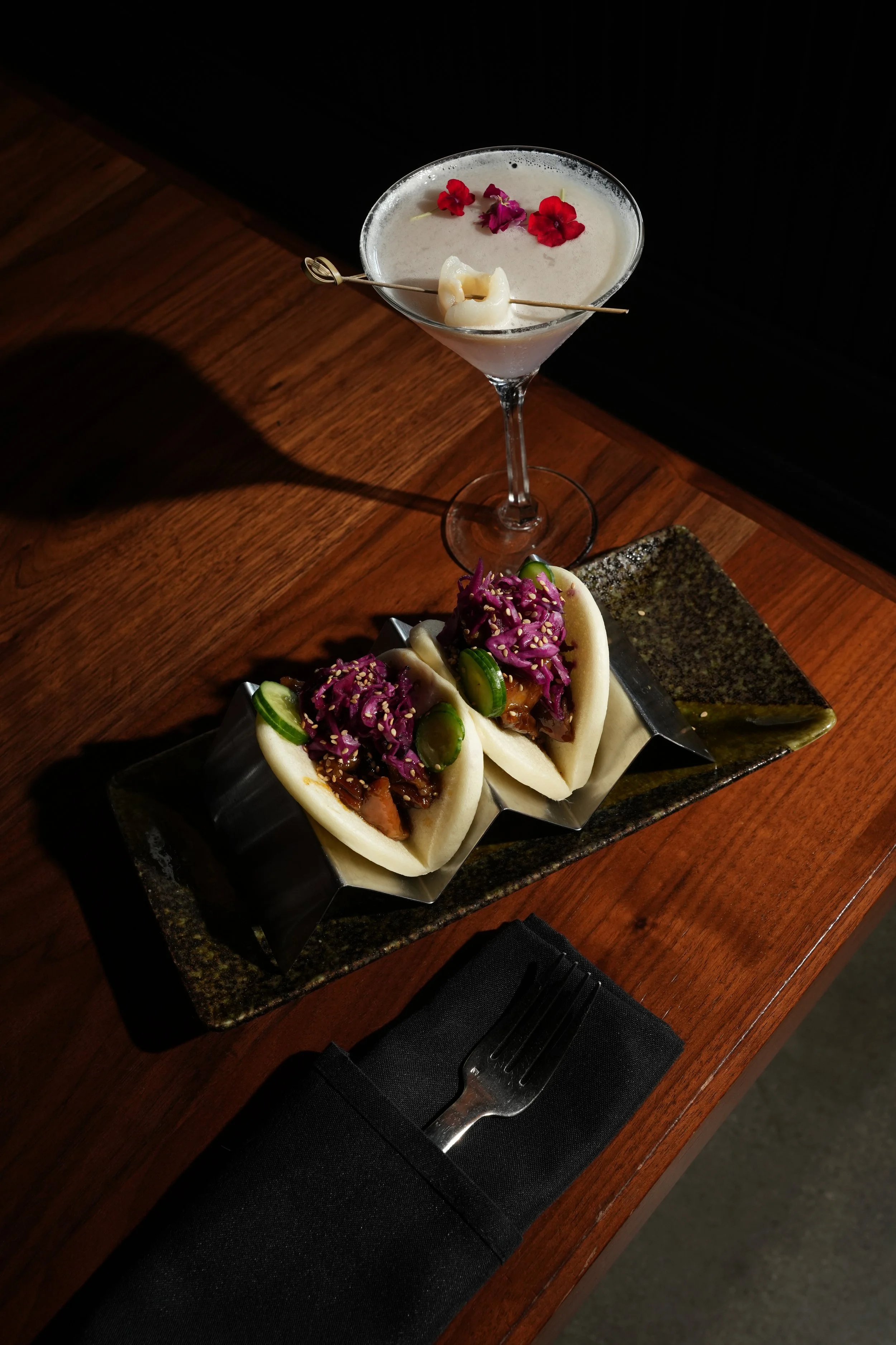 A martini glass with a white frothy drink garnished with edible flowers, a pickled vegetable, and a decorative skewer on a dark wooden table. Two boa buns with purple cabbage, cucumber slices, and sesame seeds are on a rectangular plate nearby.