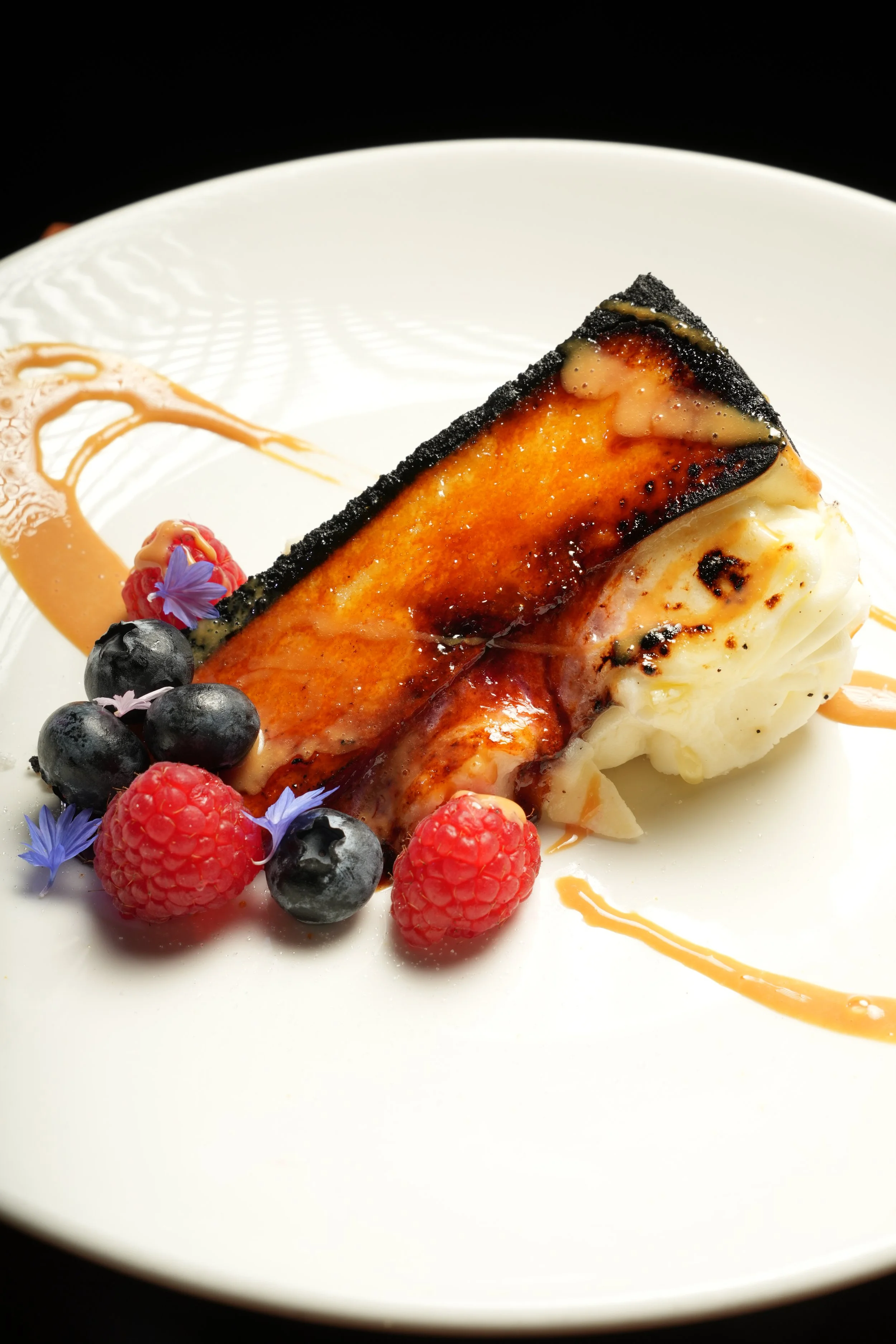A slice of cheesecake garnished with blueberries, raspberries, edible flowers, and caramel sauce on a white plate.