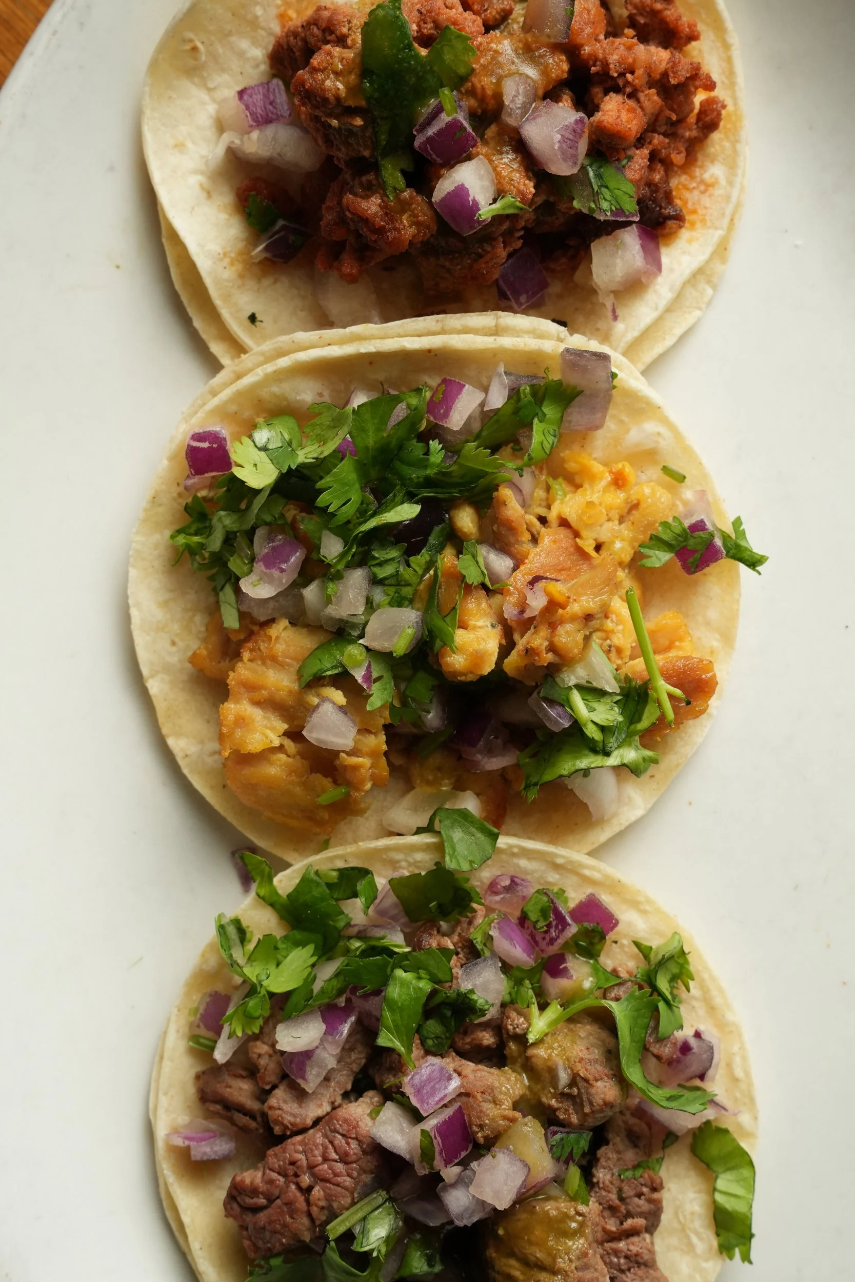Three tacos filled with seasoned beef, chopped red onions, and cilantro on a white plate. The tacos have soft corn tortillas.