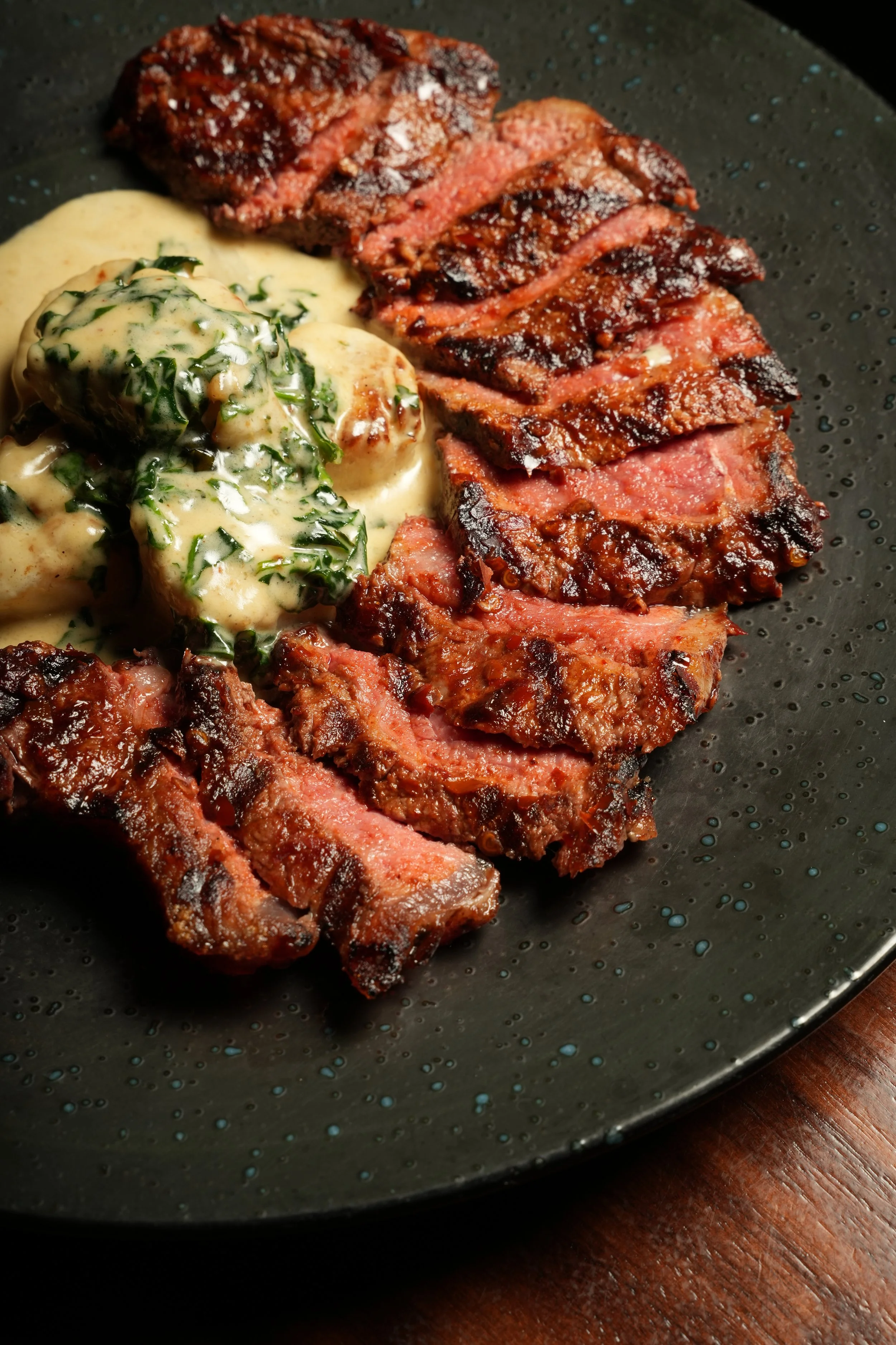 Slices of grilled steak with a slightly charred exterior and pink interior, served with a side of creamy mixture with herbs on a black textured plate.