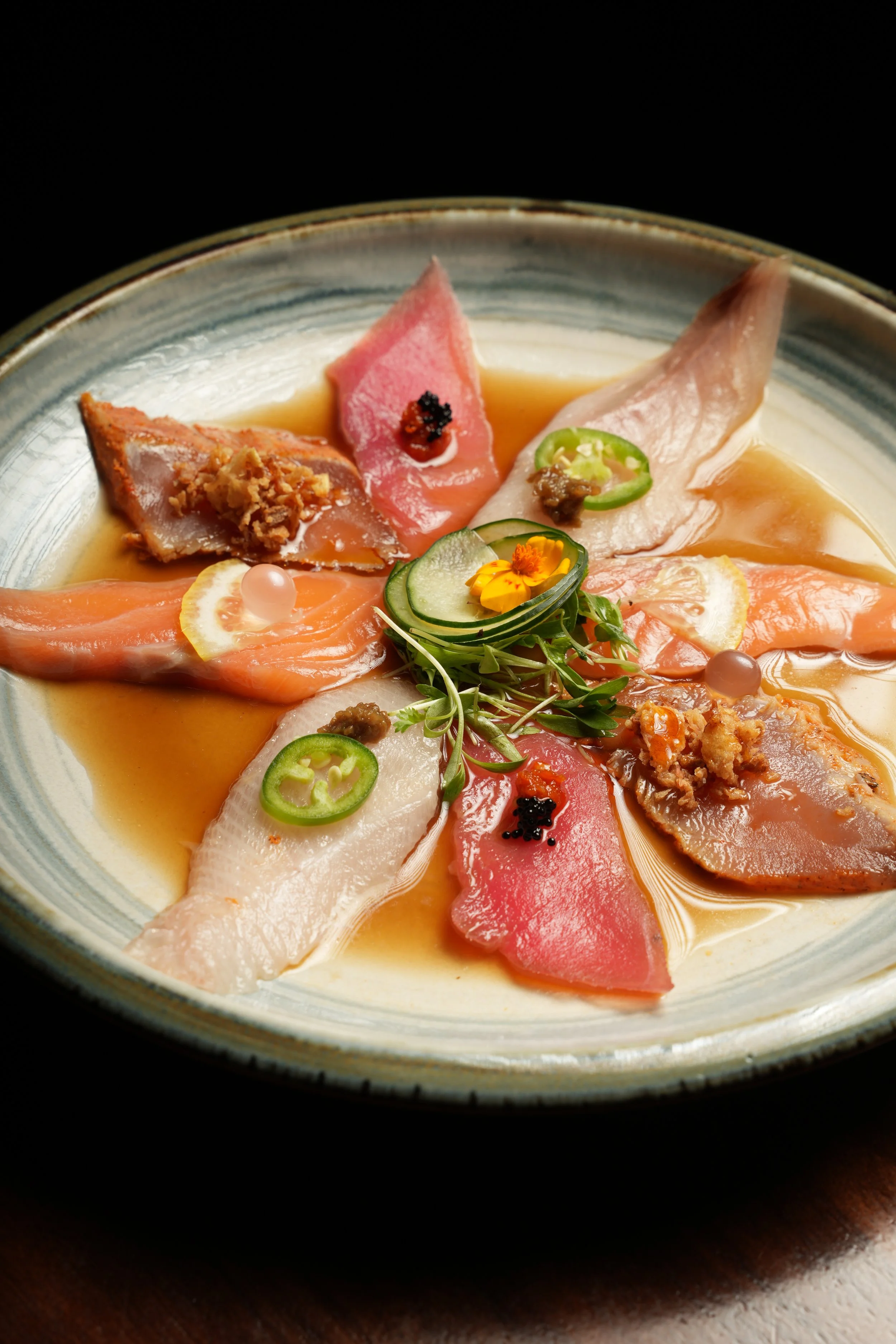 Sashimi platter with slices of raw fish, garnished with slices of lime, green chili, microgreens, and small dollops of sauce, served in a bowl with some broth.