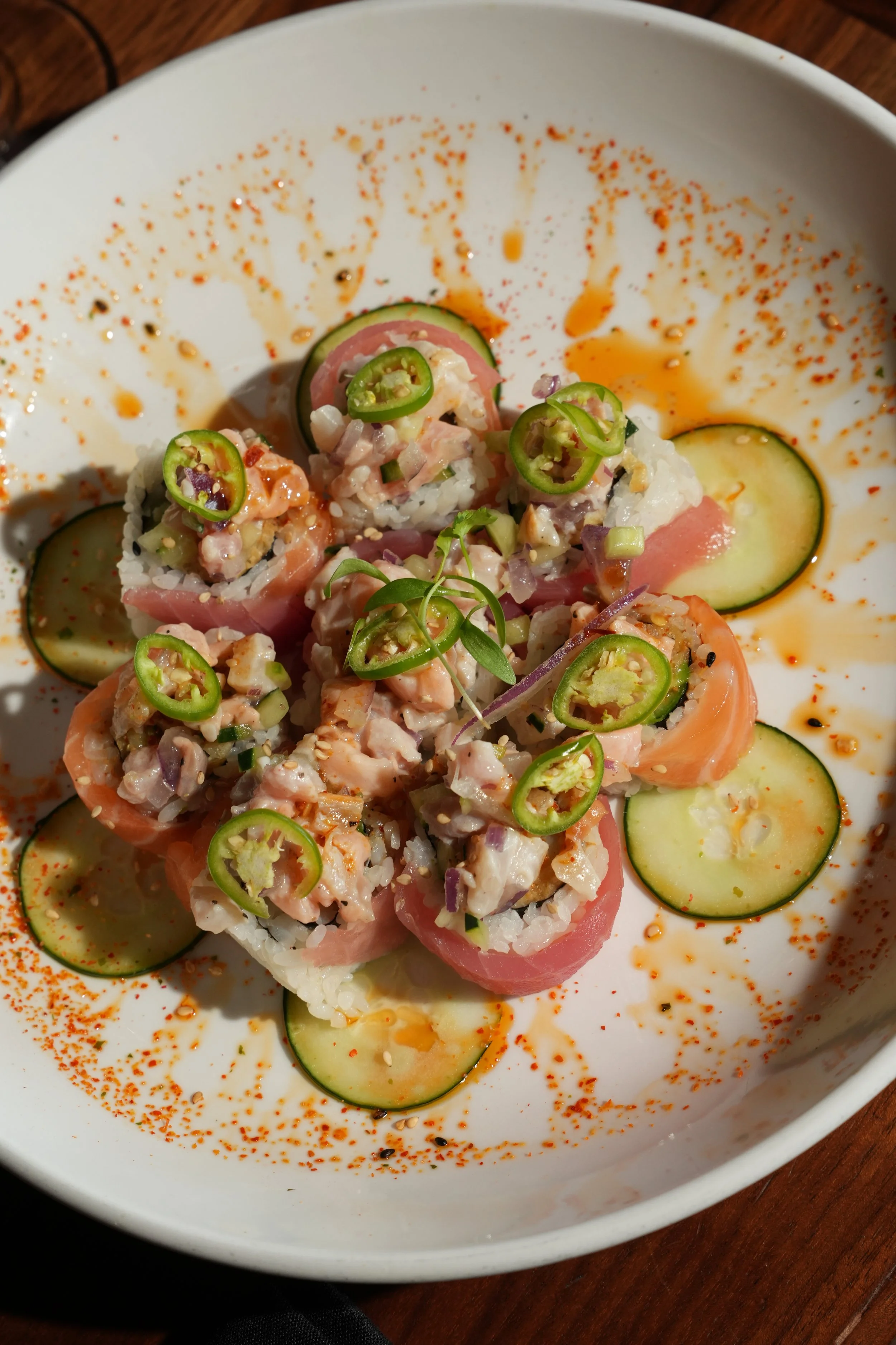 Sushi rolls topped with sliced jalapeños, surrounded by cucumber slices and sprinkled with red pepper flakes on a white plate.