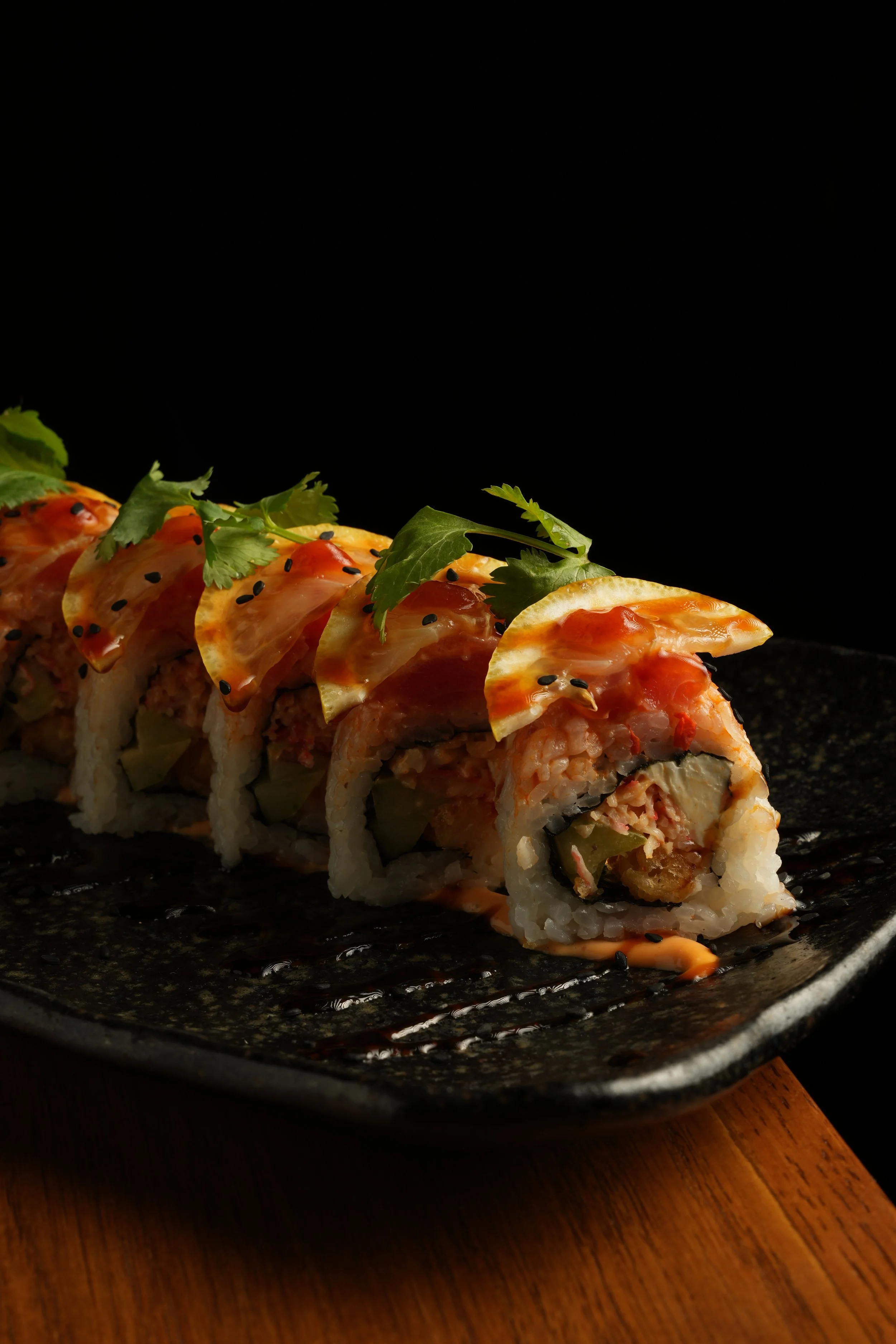 Sushi roll with rice, fish, and vegetables topped with sliced fish, sauce, cilantro, and sesame seeds, served on a black plate with a dark background