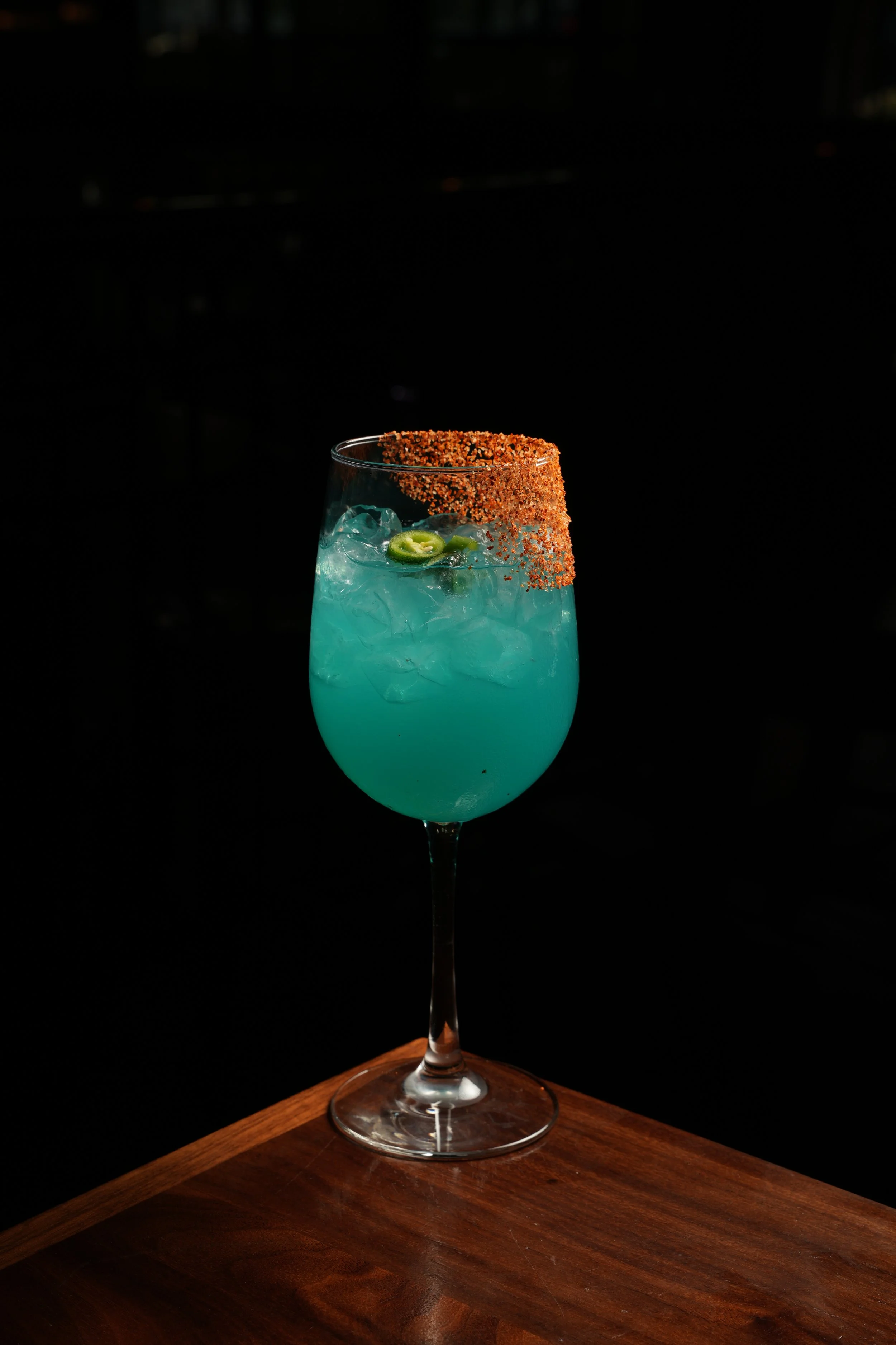 A tall wine glass filled with bright blue cocktail, garnished with green jalapeno slices and rimmed with chili powder, on a wooden surface against a black background.