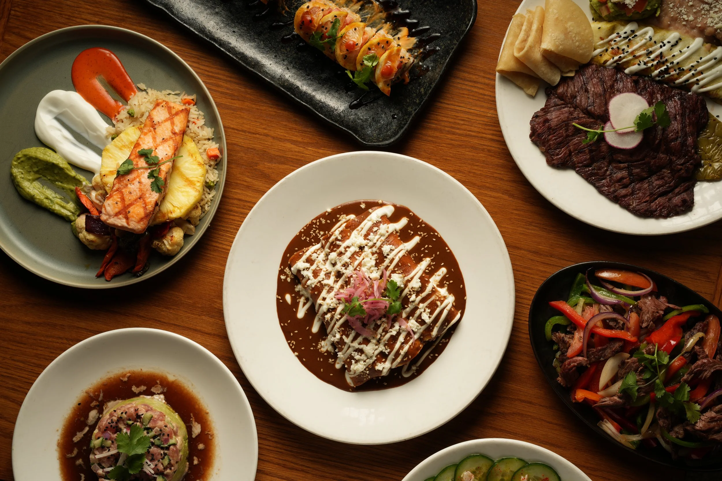 Variety of Mexican dishes on a wooden table, including grilled fish, beef steak, stuffed peppers, guacamole, and salads.
