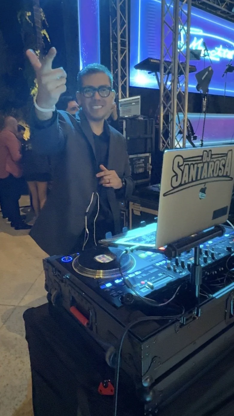 A DJ wearing glasses, smiling and pointing at the camera, standing behind a DJ setup with turntables and a laptop with a sticker that says "DJ Santarosa" at an outdoor event with blue neon lighting and a large digital screen in the background.