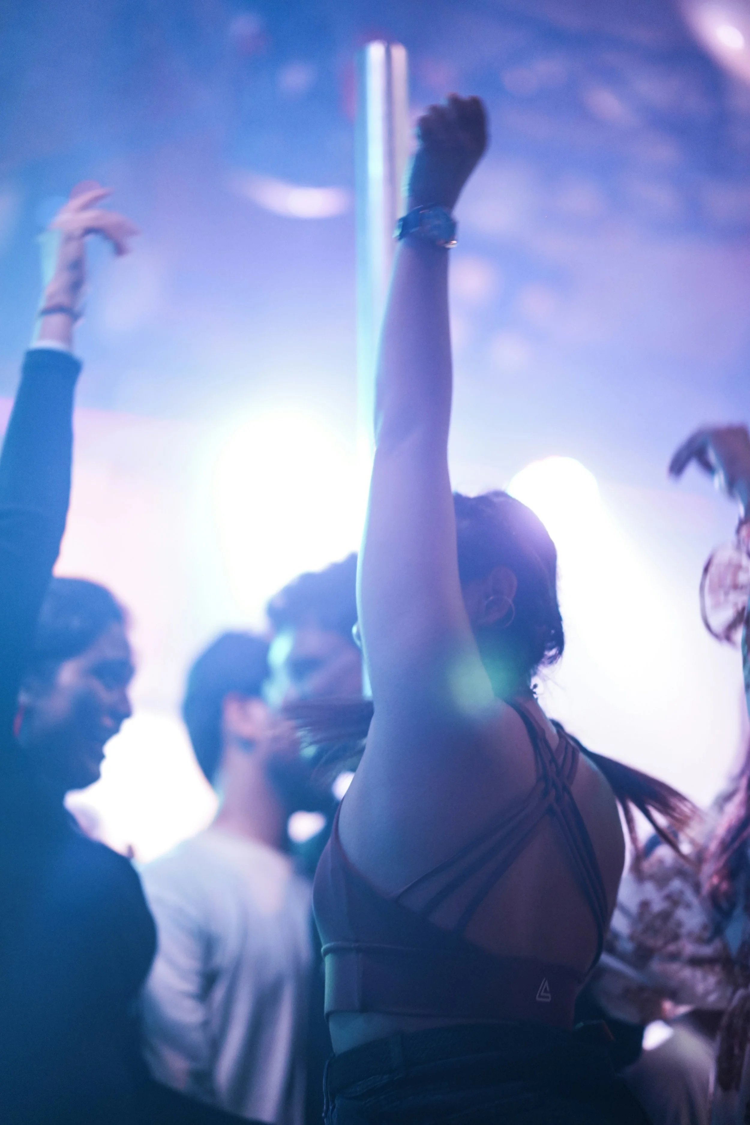 A woman dancing at a club or concert with her hand raised, wearing a sports bra and dark high-waisted pants, with colorful party lights and other people in the background.