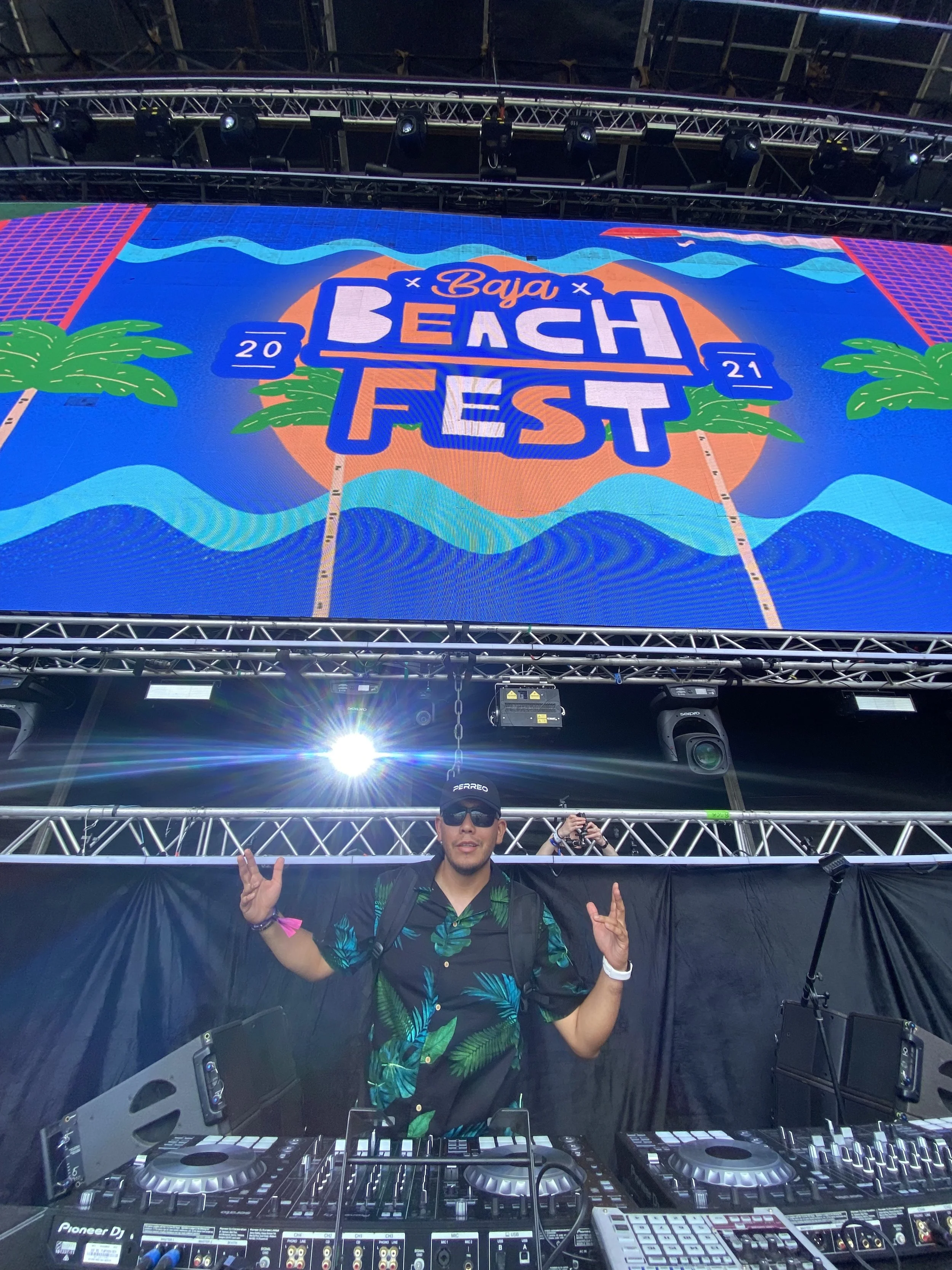 A DJ is standing behind a DJ booth at Baja Beach Fest, wearing sunglasses and a black Hawaiian shirt with green palm leaf patterns. The DJ is posing with his hands making rock and peace signs. The booth is equipped with professional DJ equipment, and large LED screens display the event's colorful logo and tropical graphics, including palm trees and waves.