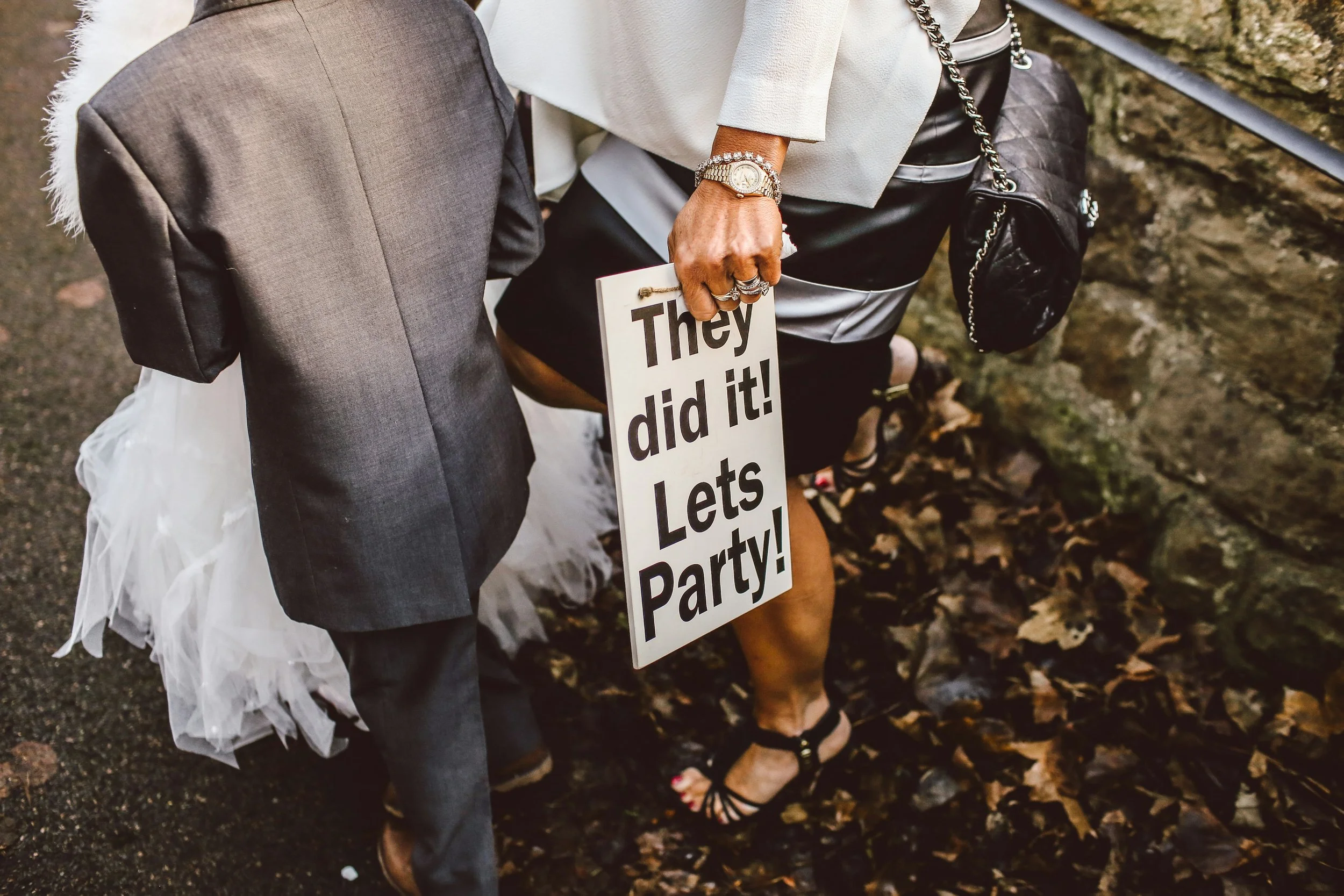 Close-up of two people dressed in formal attire, holding a sign that says "They did it! Let's Party!" at an outdoor event with fallen leaves on the ground.