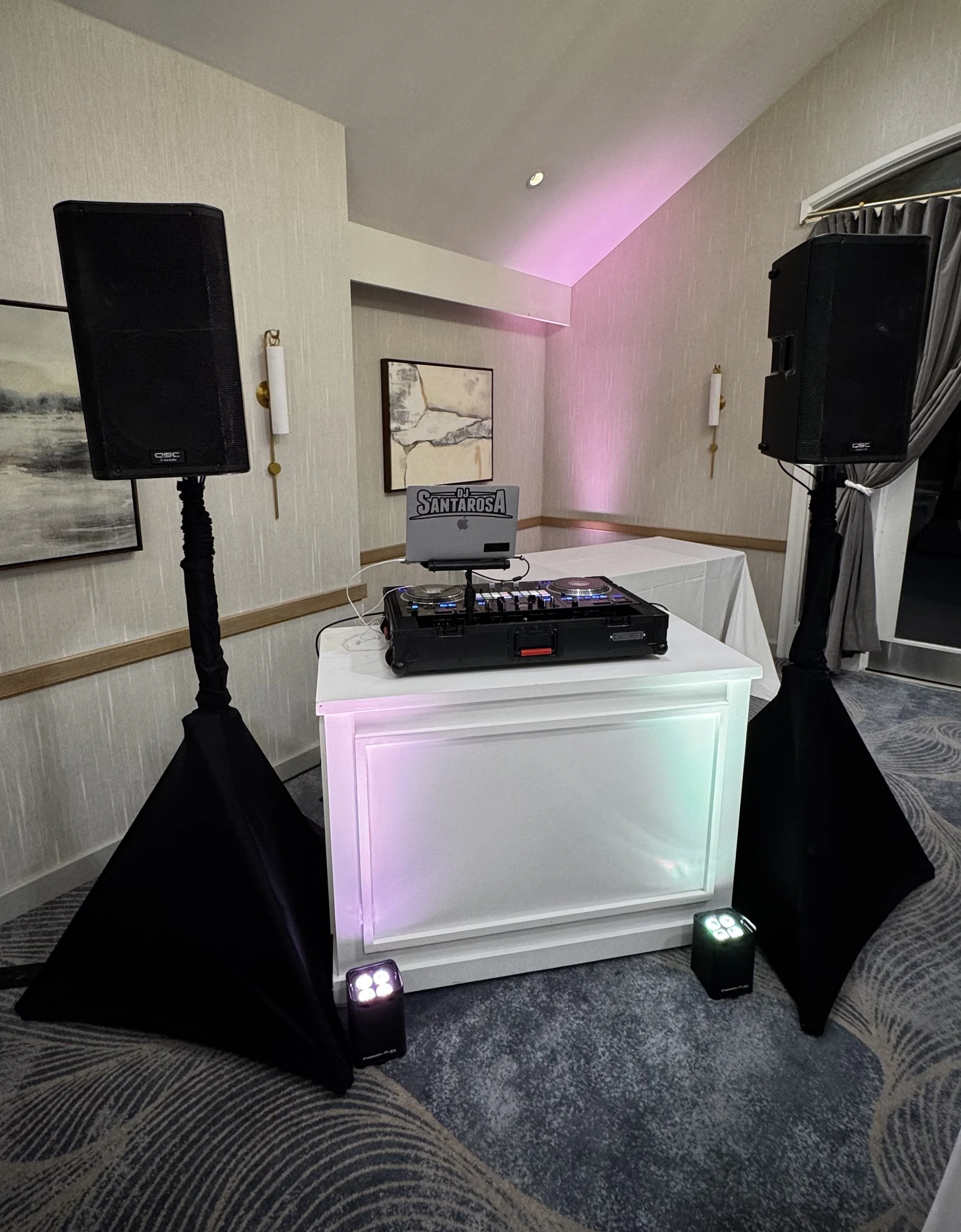A DJ setup with two large speakers on stands, a DJ console with a laptop labeled 'DJ SANTAROSA,' and colorful lighting at an indoor event space. There is a framed painting on the wall and a curtain near a window.