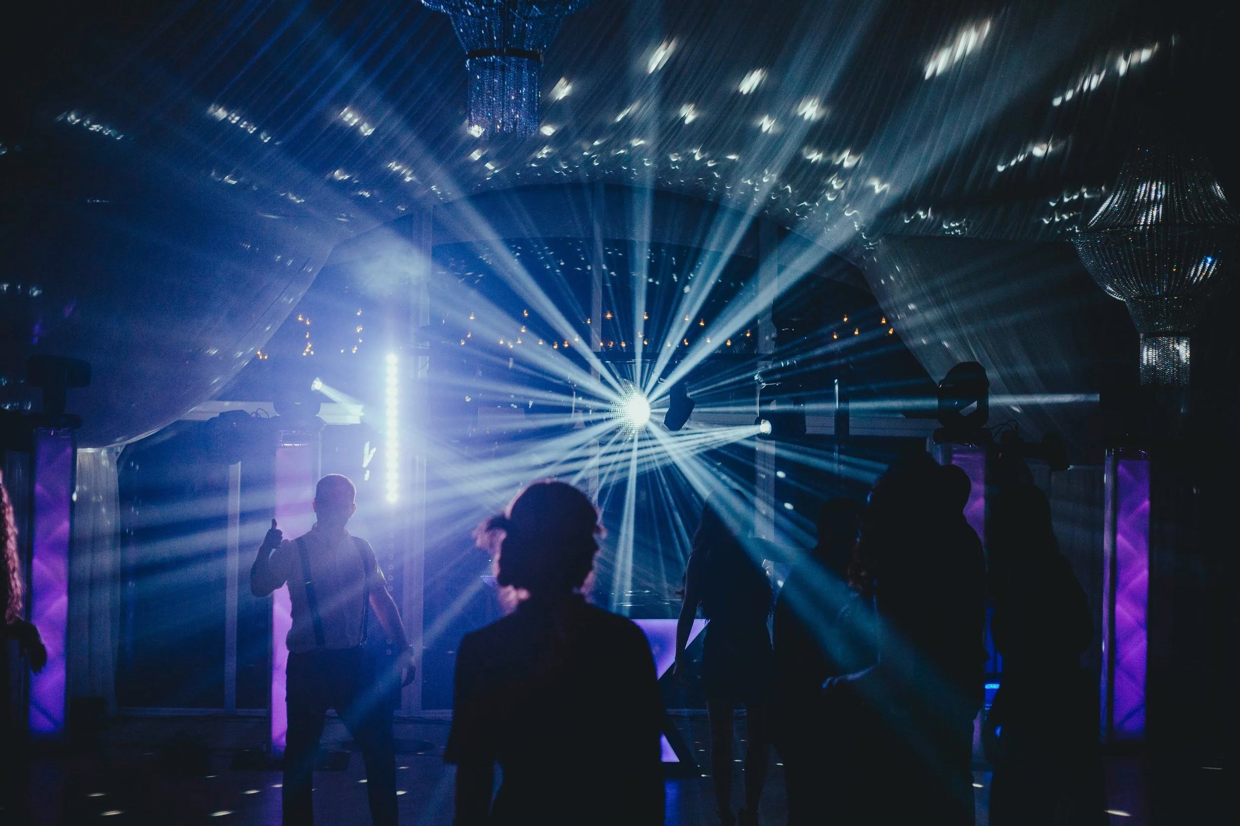 People dancing in a dimly lit nightclub with blue and purple stage lights and laser beams.