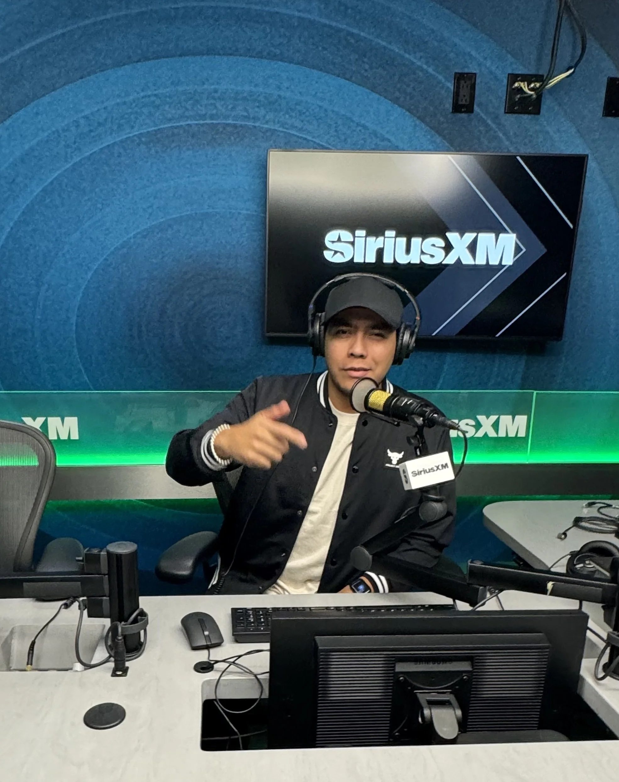 A man wearing headphones and a sports jacket sitting at a desk with computer monitors, microphone, and office supplies, in a studio with SiriusXM signage in the background.