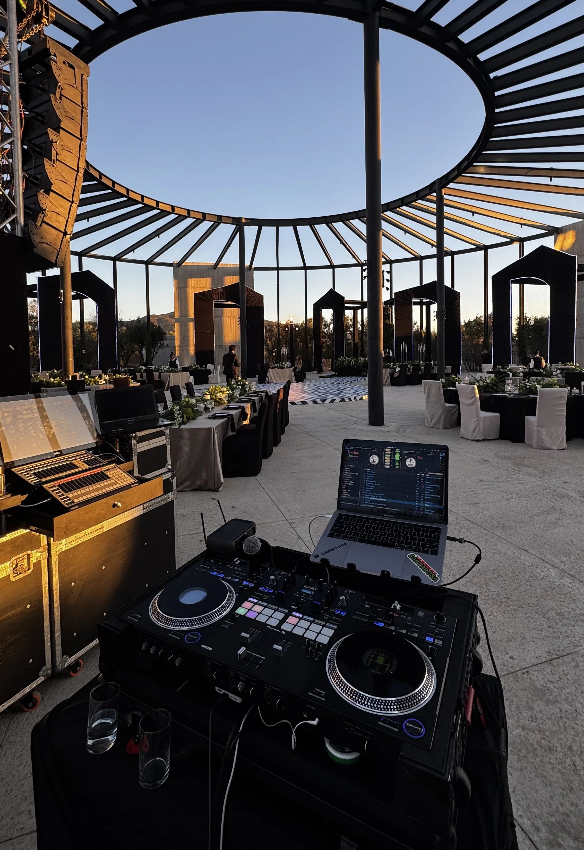 An outdoor event setup with decorated tables, chairs, and large black frames, under a circular metal structure with a view of the sunset, with DJ equipment and a laptop in the foreground.