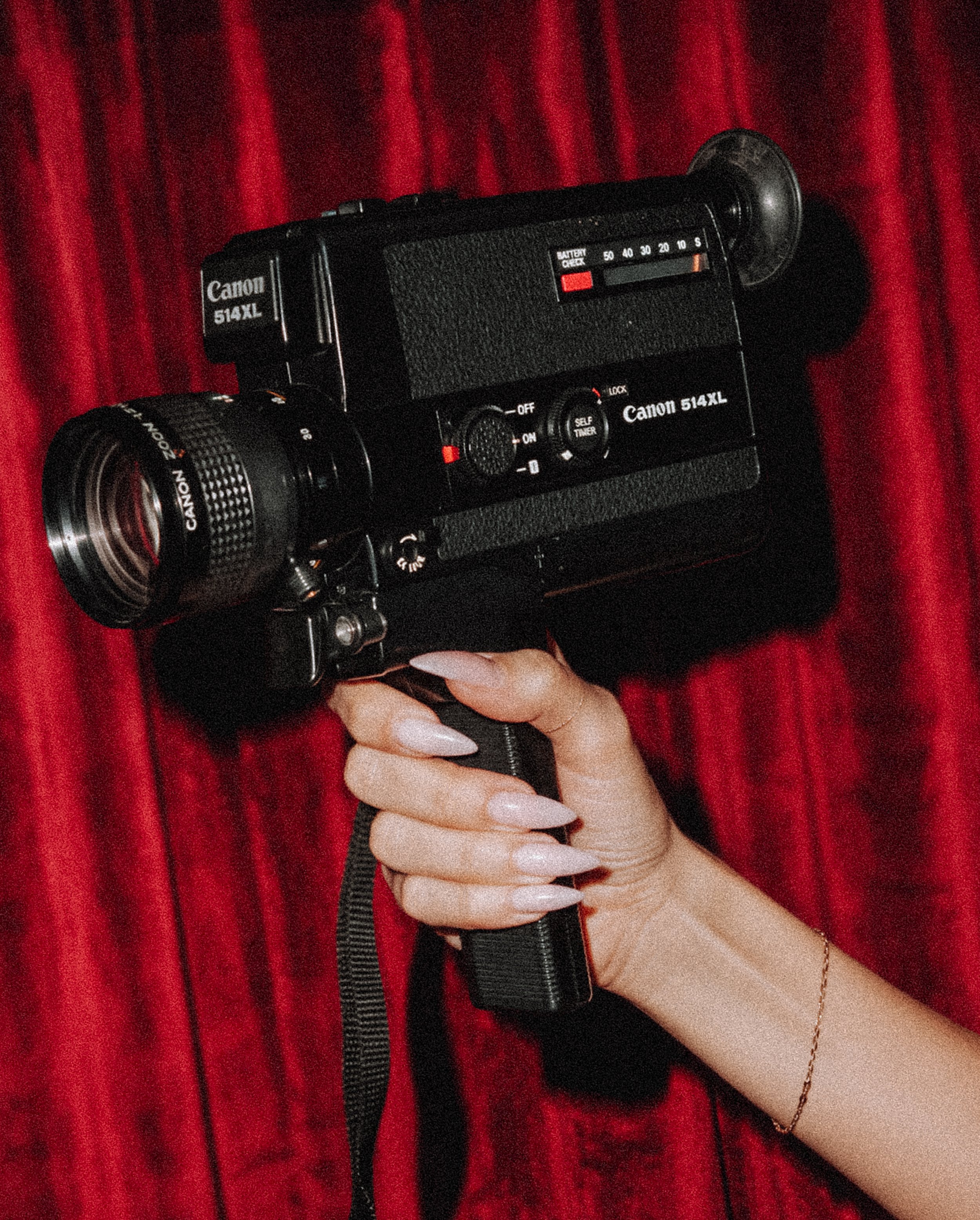 A hand holding a vintage Canon 514XL super 8 camera in front of a red curtain background.