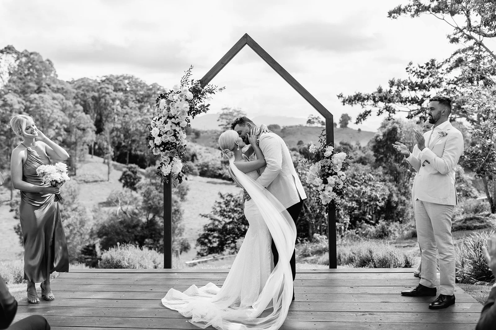 A couple kissing at their outdoor wedding ceremony under a wooden arch decorated with flowers, with bridesmaids and groomsmen clapping and smiling at a wedding at Braeside Estate