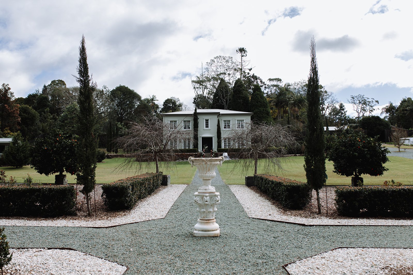 Front view of a large white house surrounded by a manicured garden with tall cypress trees, blooming and leafless trees, and a gravel pathway leading to the entrance at a wedding at Wynmere Estate.