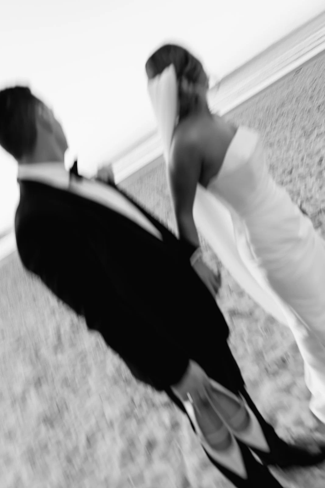 A black and white photo of a man in a tuxedo and a woman in a wedding dress standing together indoors. The woman is facing slightly away from the camera, and the man is turned toward her at a wedding at Maison La Plage.