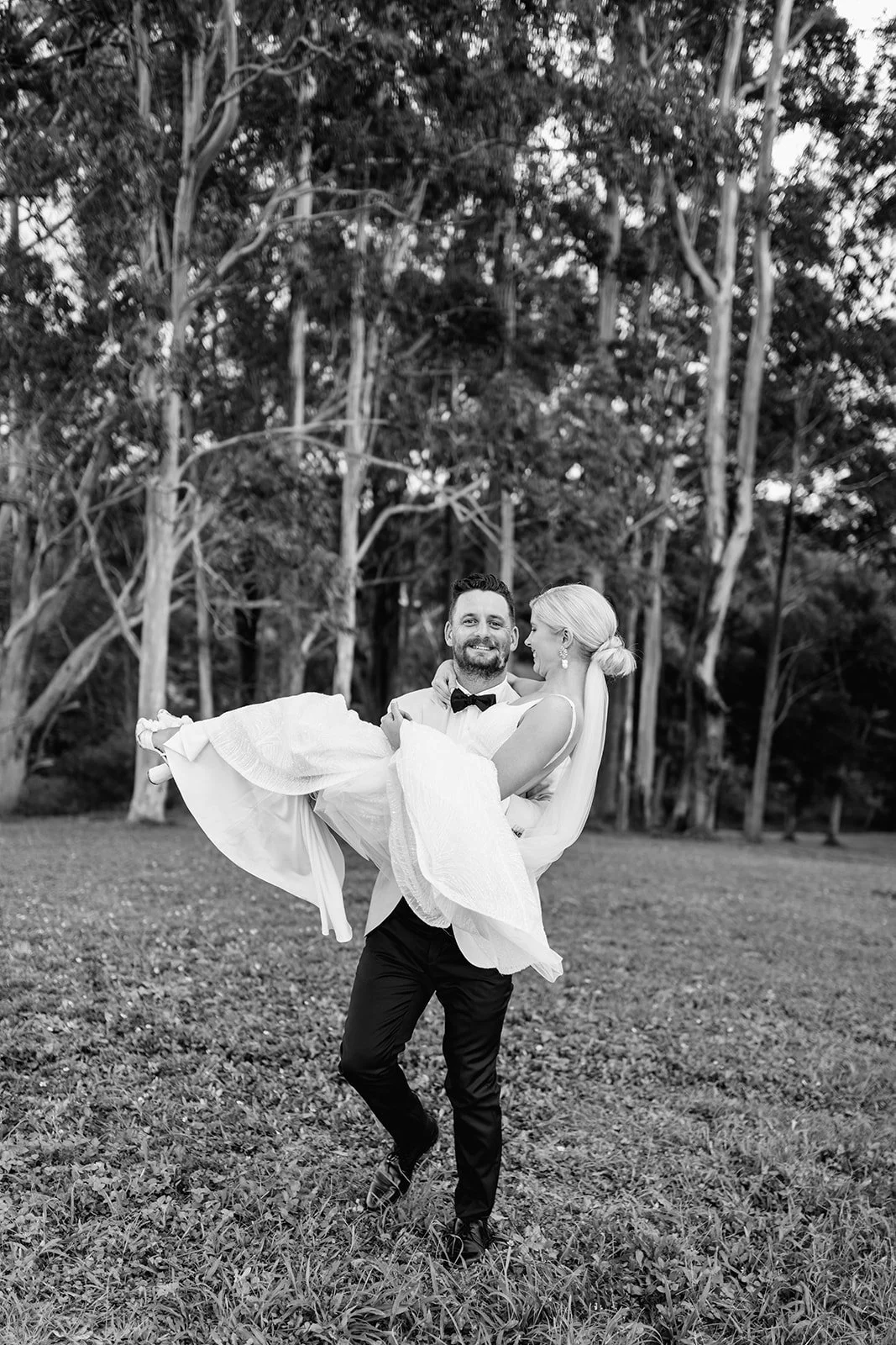 A man in a tuxedo carrying a woman in a wedding dress outdoors with trees in the background, black and white photograph at a wedding at Austinvilla Estate.