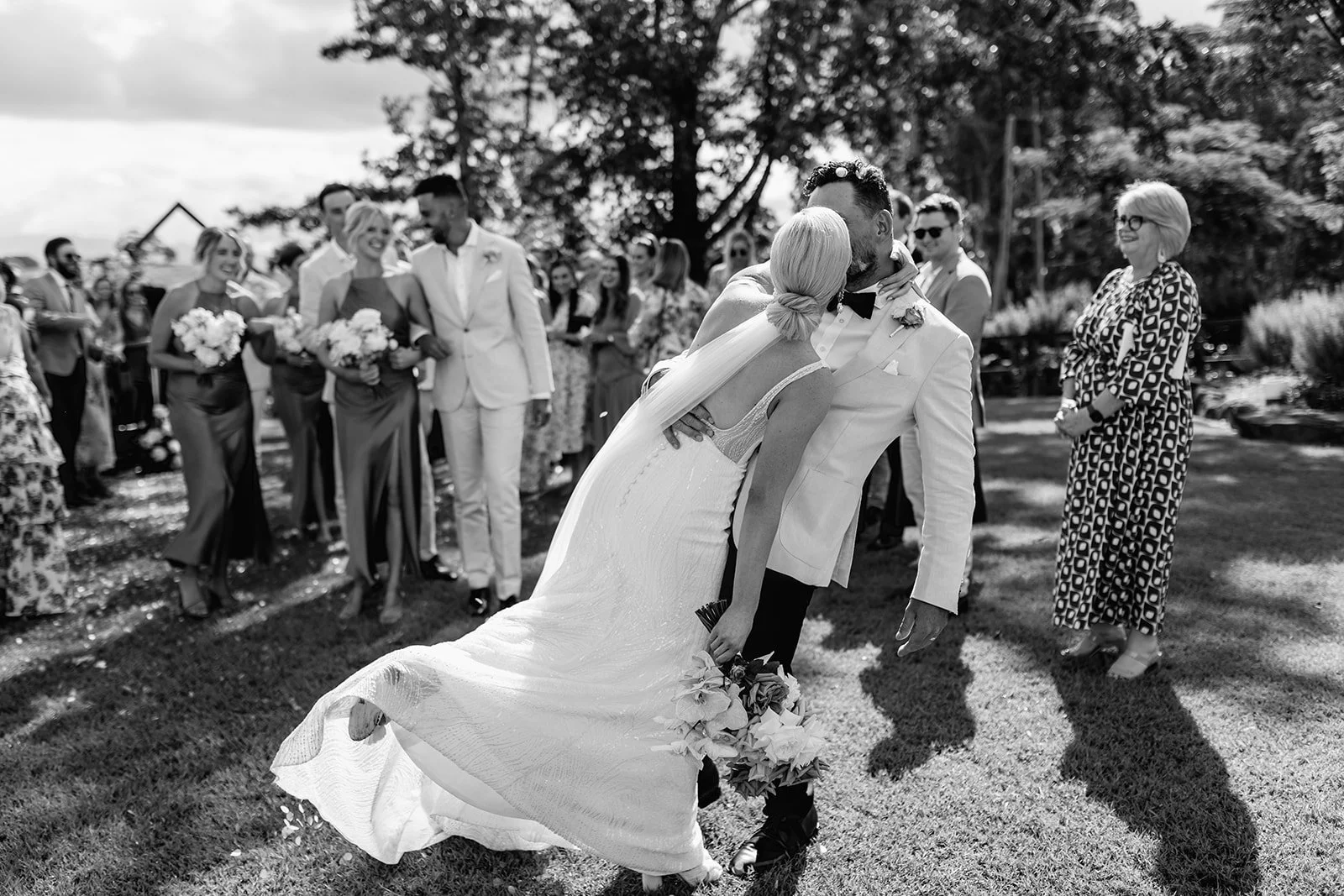 Black and white photo of a wedding scene outdoors, where a bride and groom are dancing while a group of guests and bridesmaids watch and smile in the background at Austinvilla Estate.