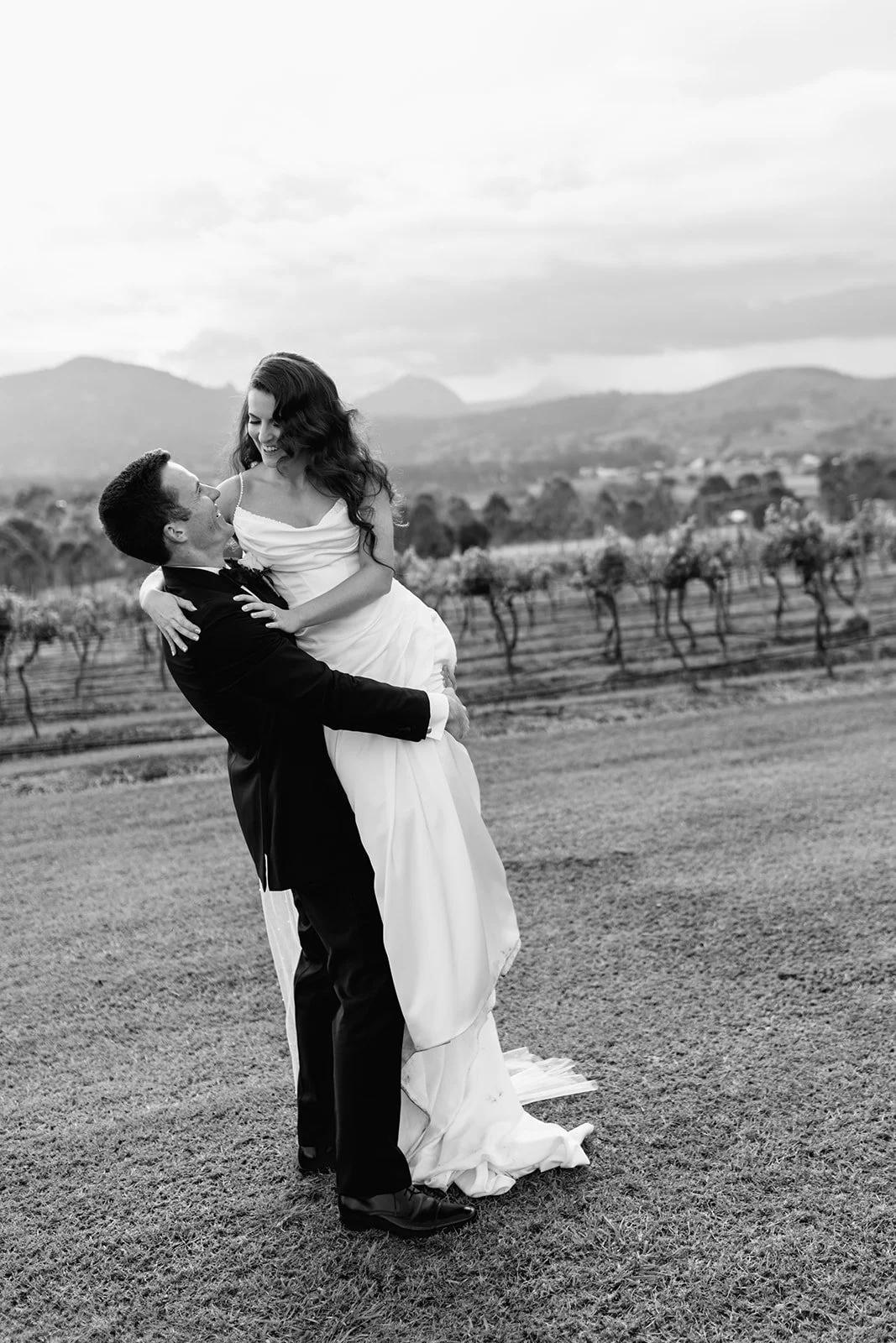 A black-and-white photo of a man lifting a woman in a wedding dress outdoors with mountains and vineyards in the background at kooroomba Lavender Farm.