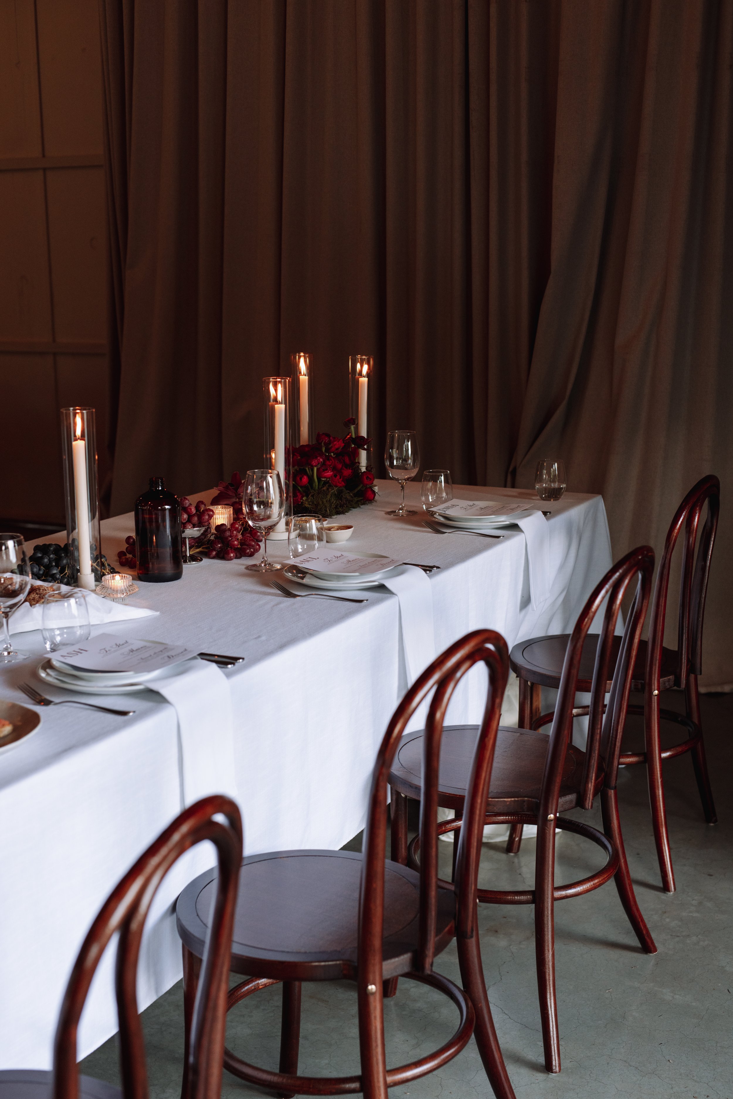 An elegant dining table set for a meal with white napkins, wine glasses, plates, and silverware, decorated with red roses, candles, and a dark bottle, in a room with wooden walls and curtains at a wedding at Frida's Field in Byron Bay.