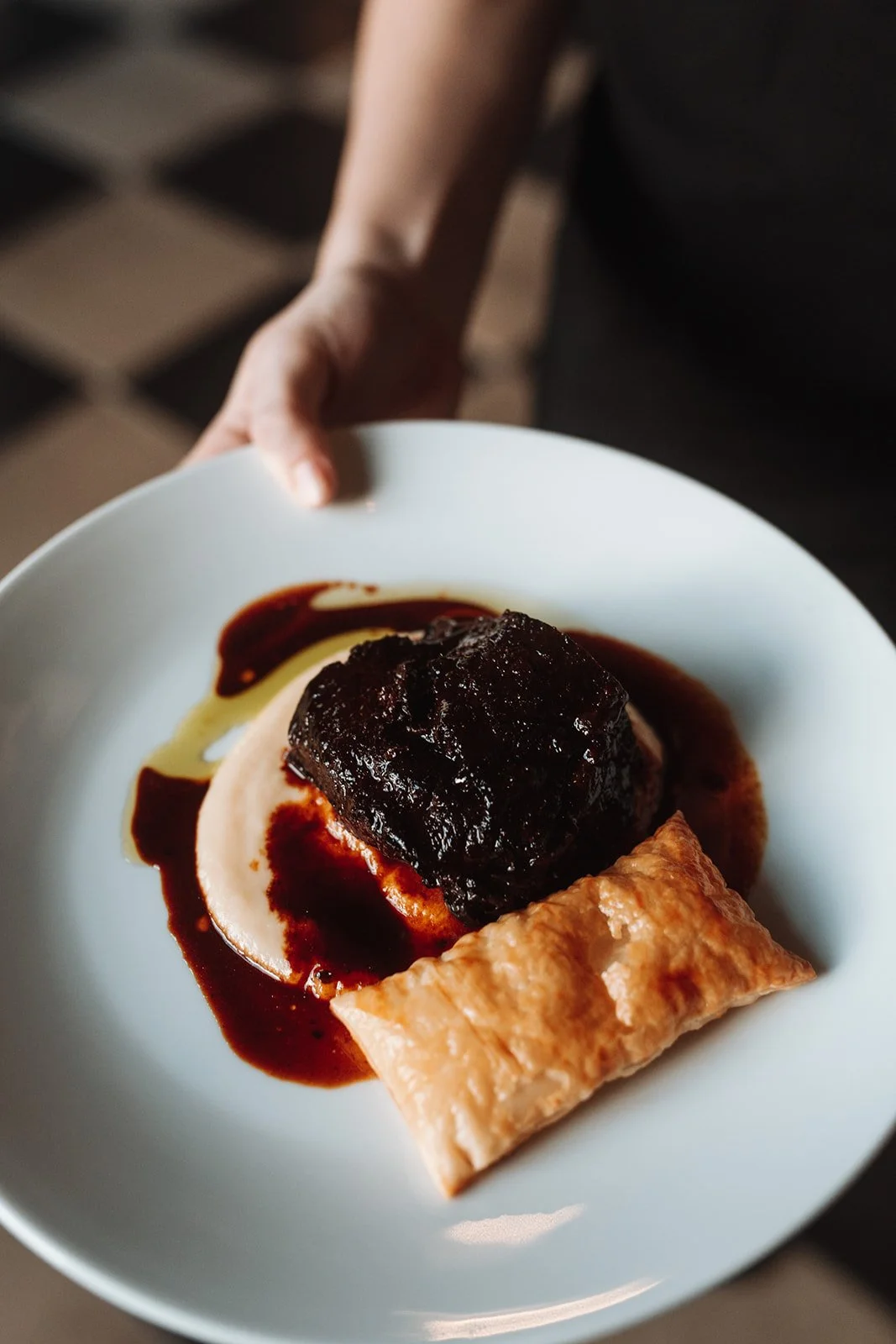 A person holding a white plate with a piece of cooked meat topped with dark sauce, a puff pastry, and a base of creamy sauce with garnish sauce around it. Fine Dining at a wedding at Campbell point House.