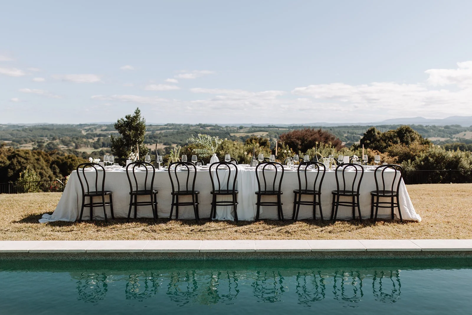 Long outdoor dining table set up with white tablecloth, glassware, and chairs, overlooking a scenic landscape with trees and hills under a partly cloudy sky, beside a swimming pool. A beautiful Wedding at the Temple Farmhouse