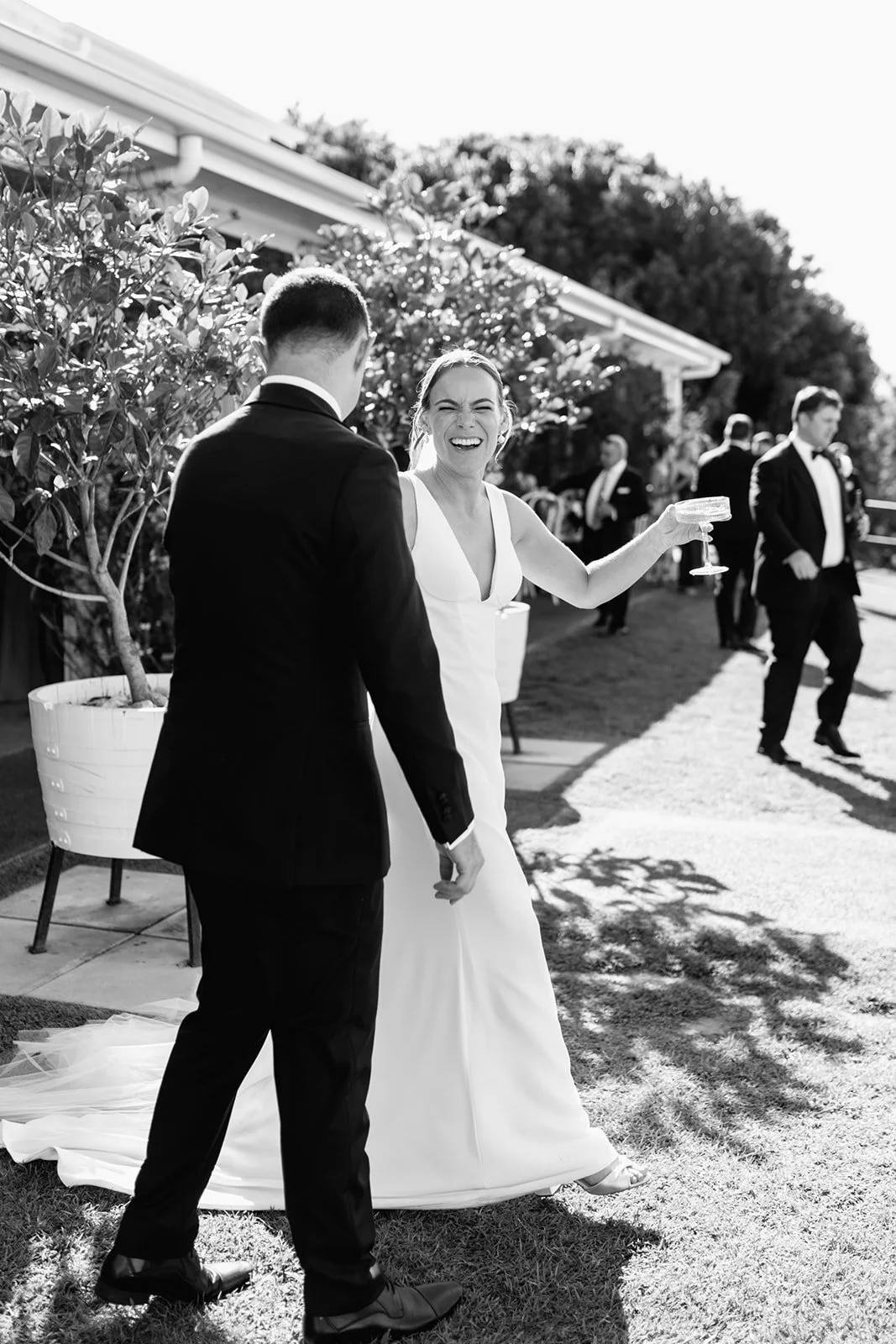 Black and white photo of a woman in a white dress holding a champagne glass, smiling and sharing a moment with a man in a tuxedo at an outdoor wedding reception at Summergrove Estate.