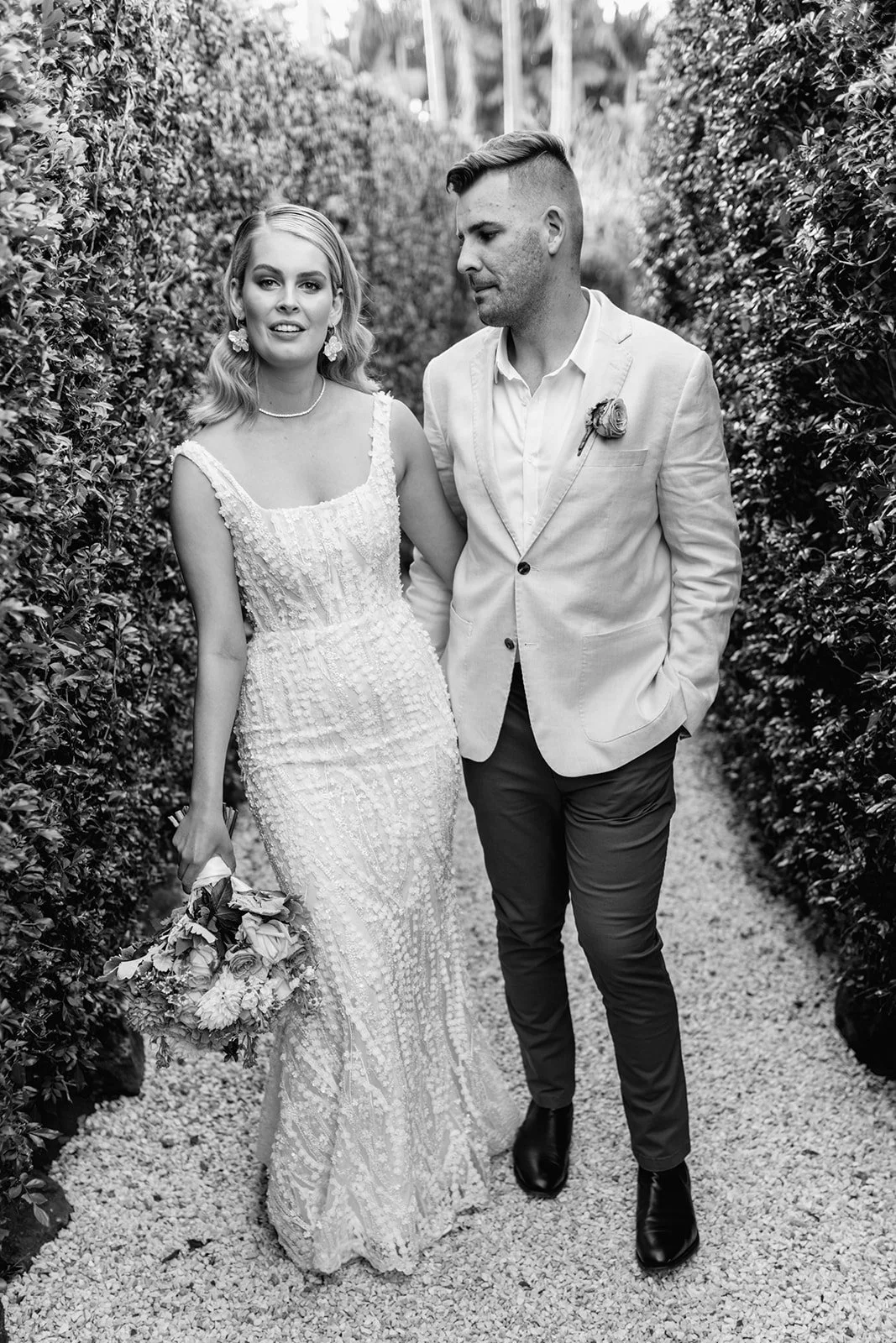 A black and white photograph of a bride and groom walking together outdoors through a hedge-lined path; the bride is wearing a detailed lace wedding dress, holding a bouquet, and the groom is in a suit at a wedding at Figtree Restaurant.