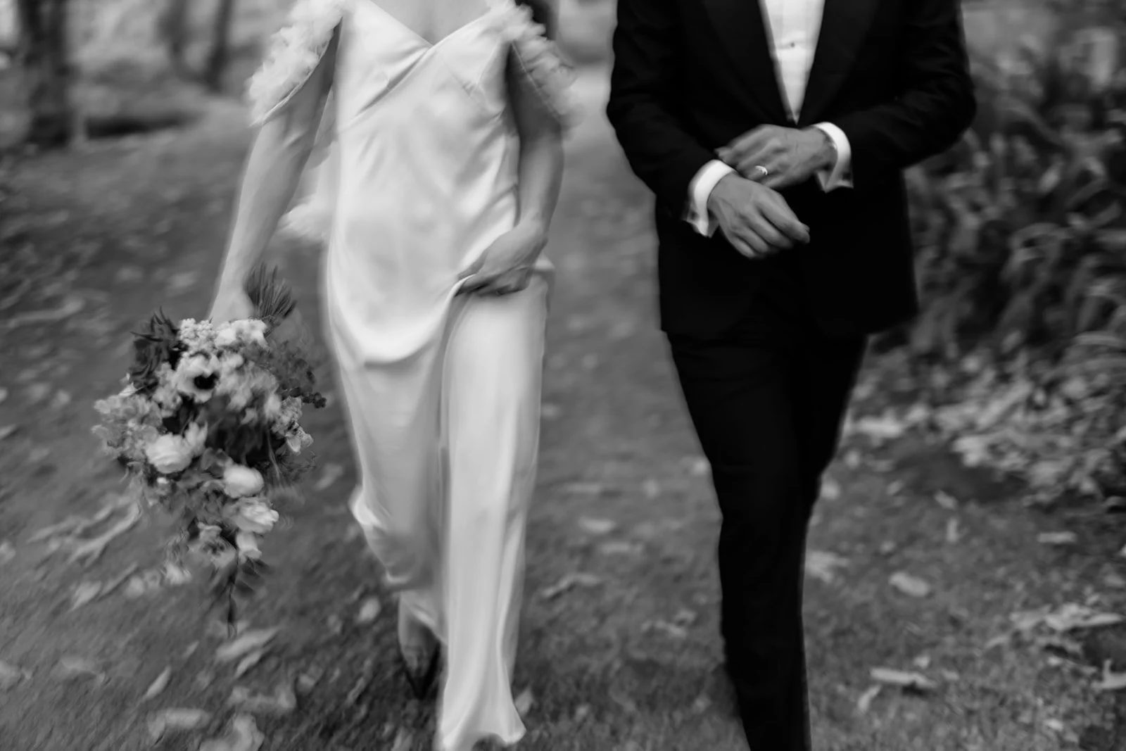 A black and white photo of a couple walking outdoors, the woman in a Silk Laundry wedding dress holding a bouquet, and the man in a suit.