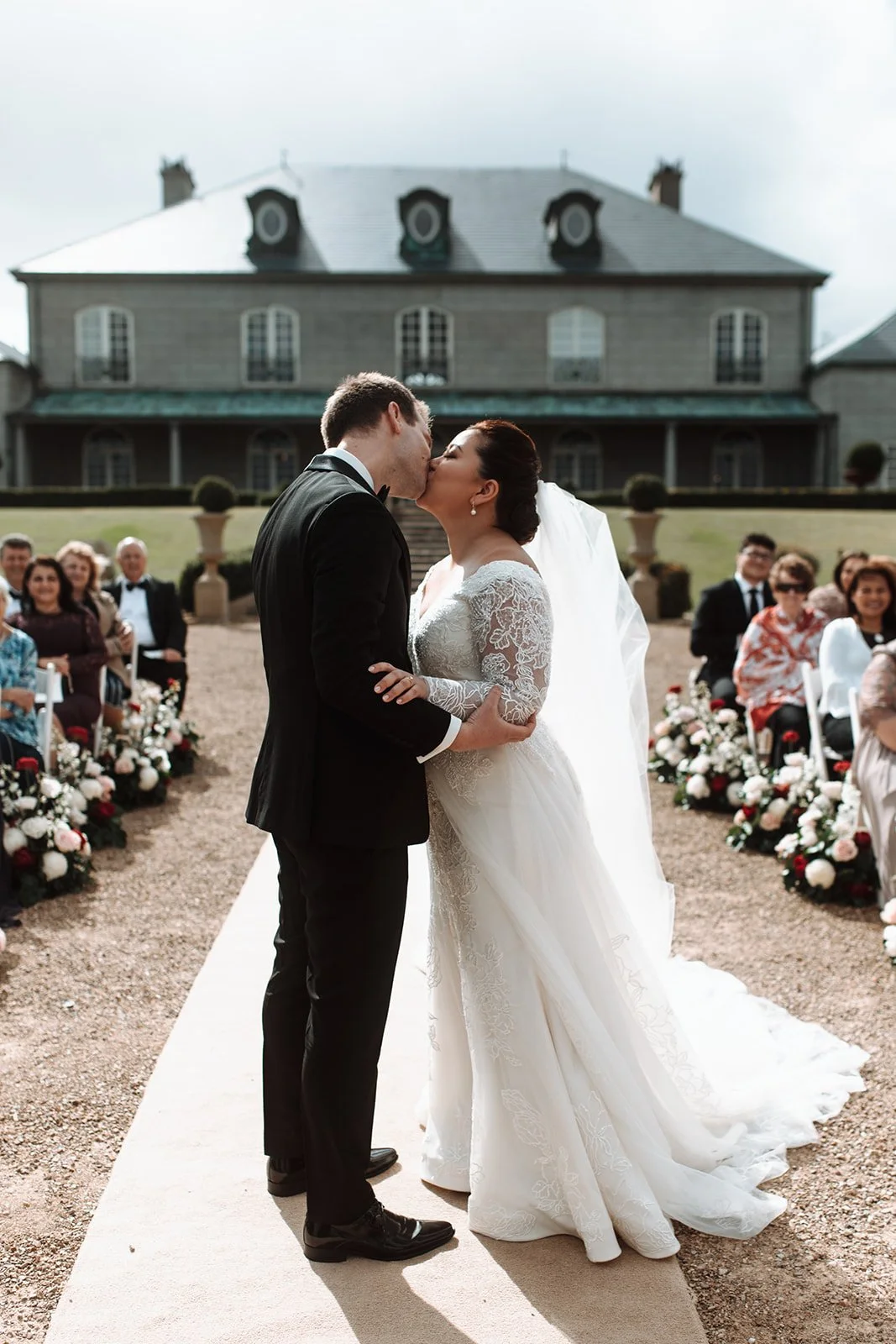 A bride and groom kiss during their outdoor wedding ceremony in front of a large house, with guests seated on either side and floral arrangements along the aisle at Campbell Point House
