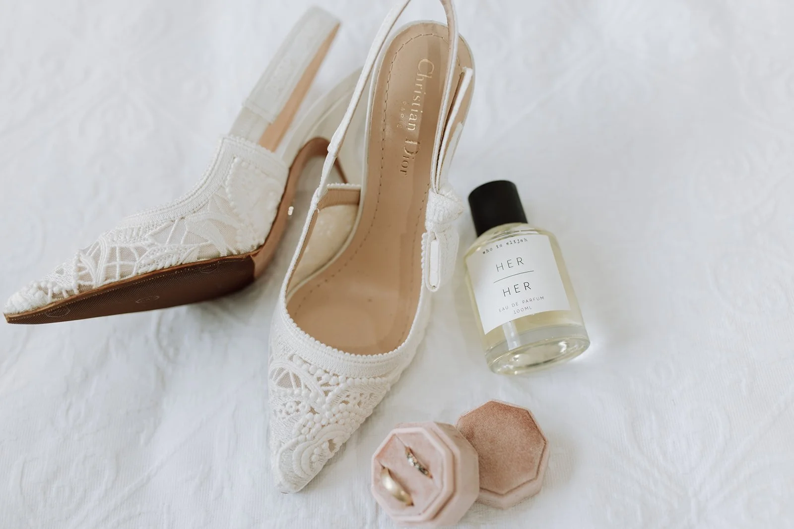 A pair of Christian Dior Wedding shoes, a bottle of perfume labeled "Her" by Christian Dior, and two pink jewelry boxes with rings inside are arranged on a white textured surface.