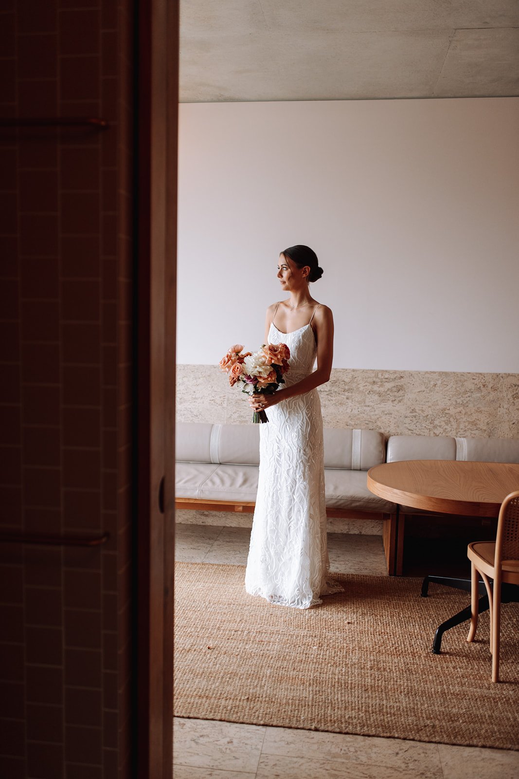A bride in a white lace dress holding a bouquet of peach, white, and pink flowers, standing in a minimalist room with a beige sofa, round wooden table, and rattan chair at a wedding at the Calile Hotel.
