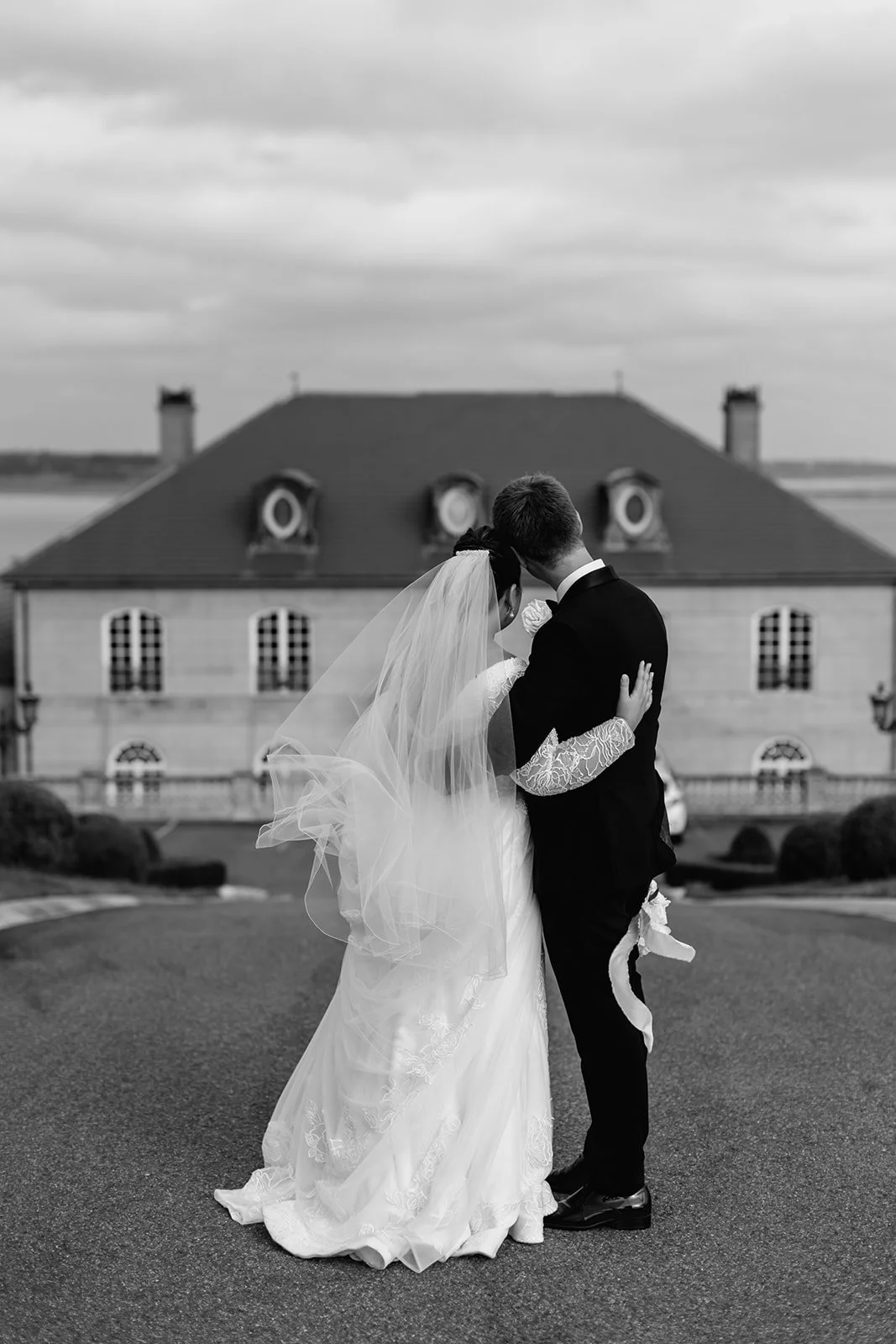 Black and white photograph of a bride and groom embracing outdoors, with a large French Chateau Wedding with multiple windows and a sloped roof in the background at Campbell Point House.