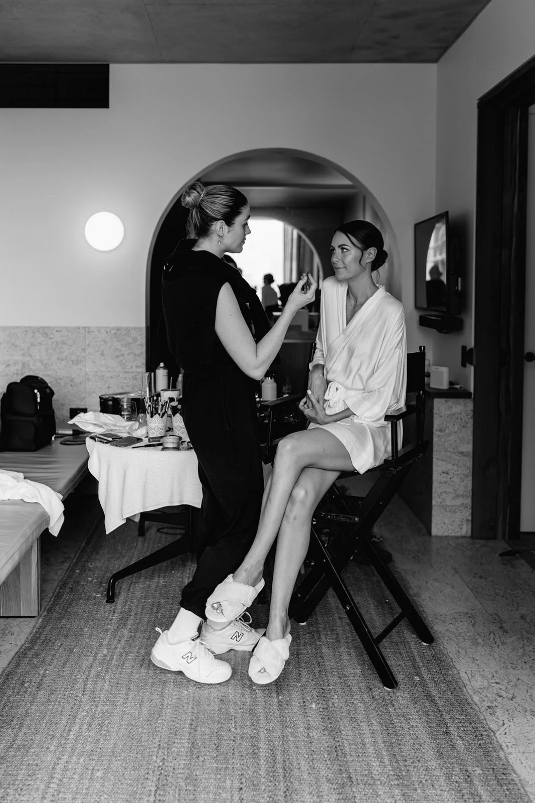 A woman in a white robe sits on a chair as another woman applies makeup or performing makeup artistry in a room with makeup supplies and a lamp, with a mirror on the wall getting ready for a wedding at the Calile Hotel.