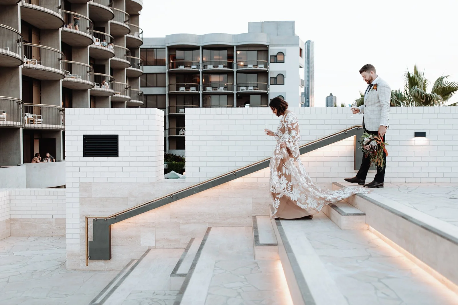 A bride and groom walking down stairs at an outdoor wedding at The Calile Hotel with modern building and palm trees in the background.