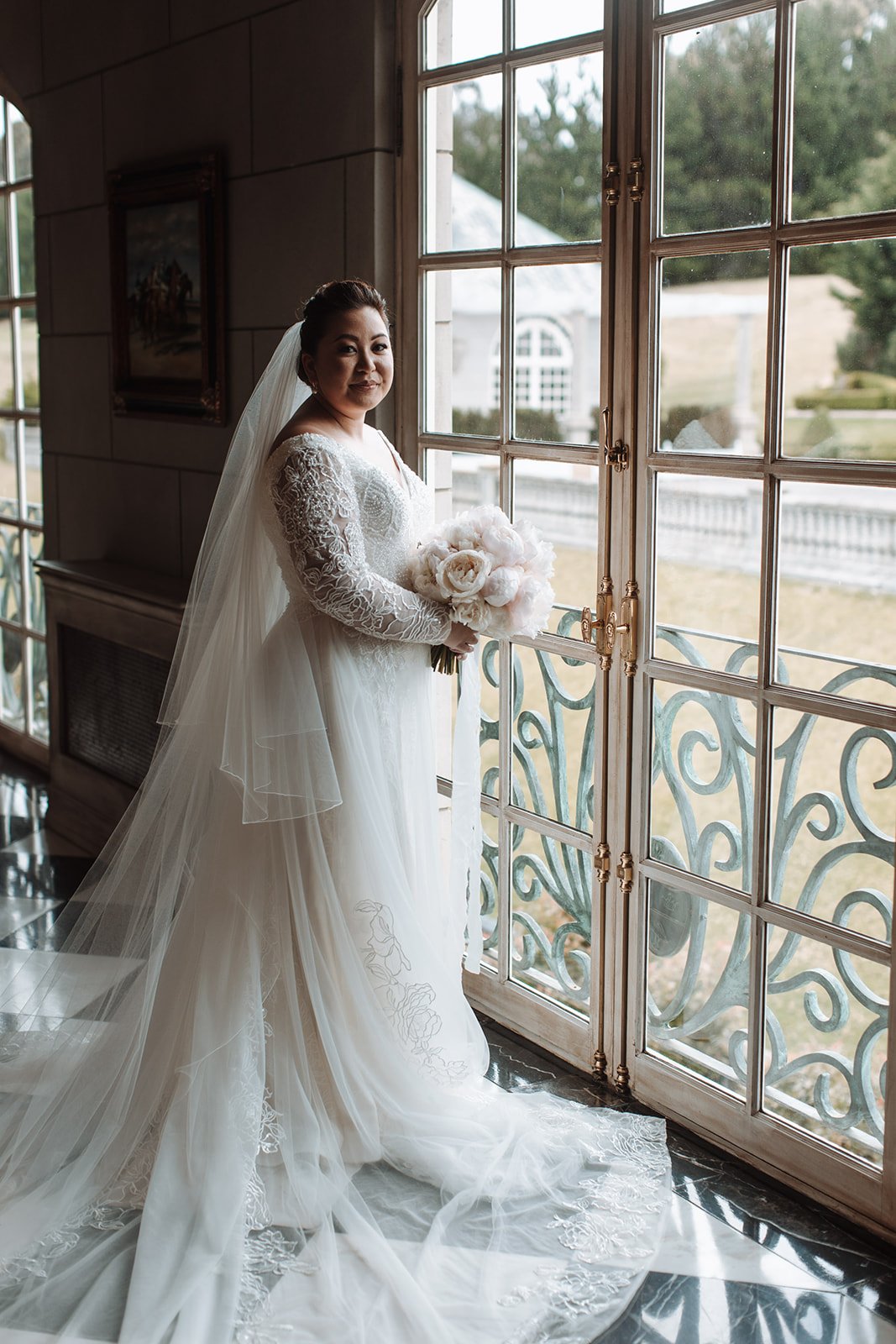 Bride in a white wedding gown with lace sleeves and a veil holding a bouquet of peonies, standing by a window with an outdoor landscape view of Campbell Point House.