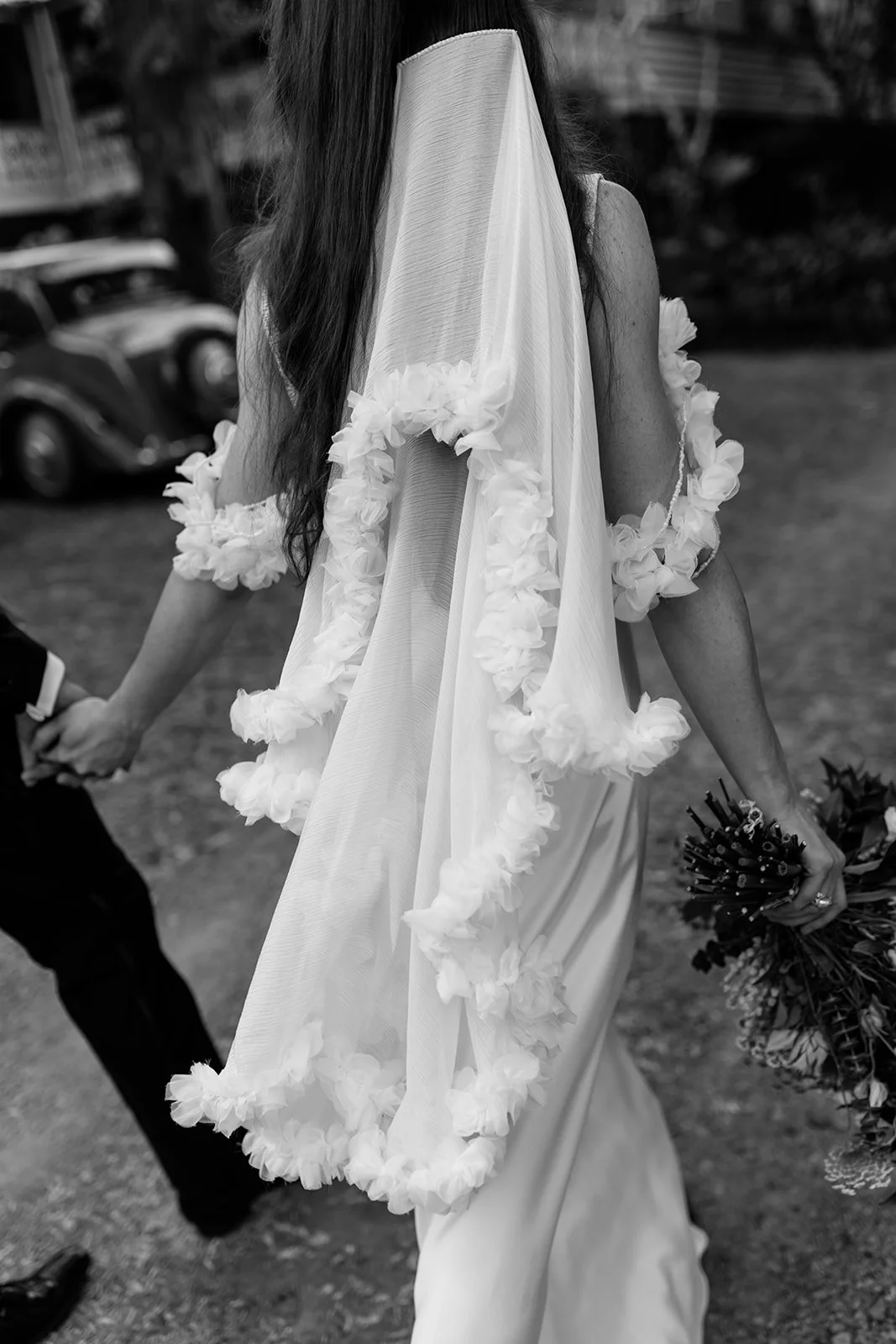 A bride in a Silk Laundry wedding dress holding a bouquet of flowers, walking outdoors, with a groom holding her hand, black and white photo at Braeside Estate
