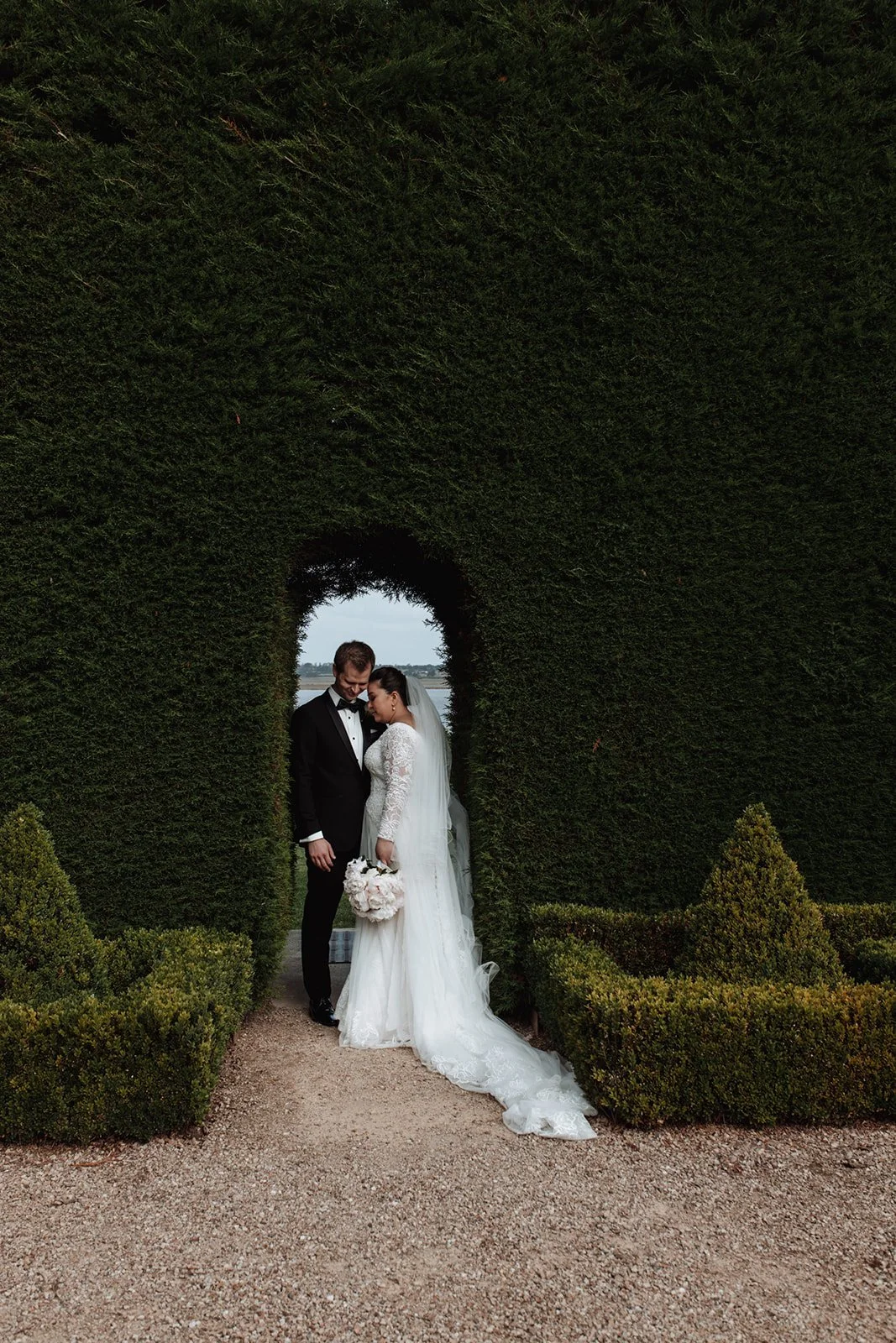 A bride and groom standing close together under a large green hedge archway, with the bride holding a bouquet of white flowers, outdoors with water visible in the distance at a wedding at Campbell Point House